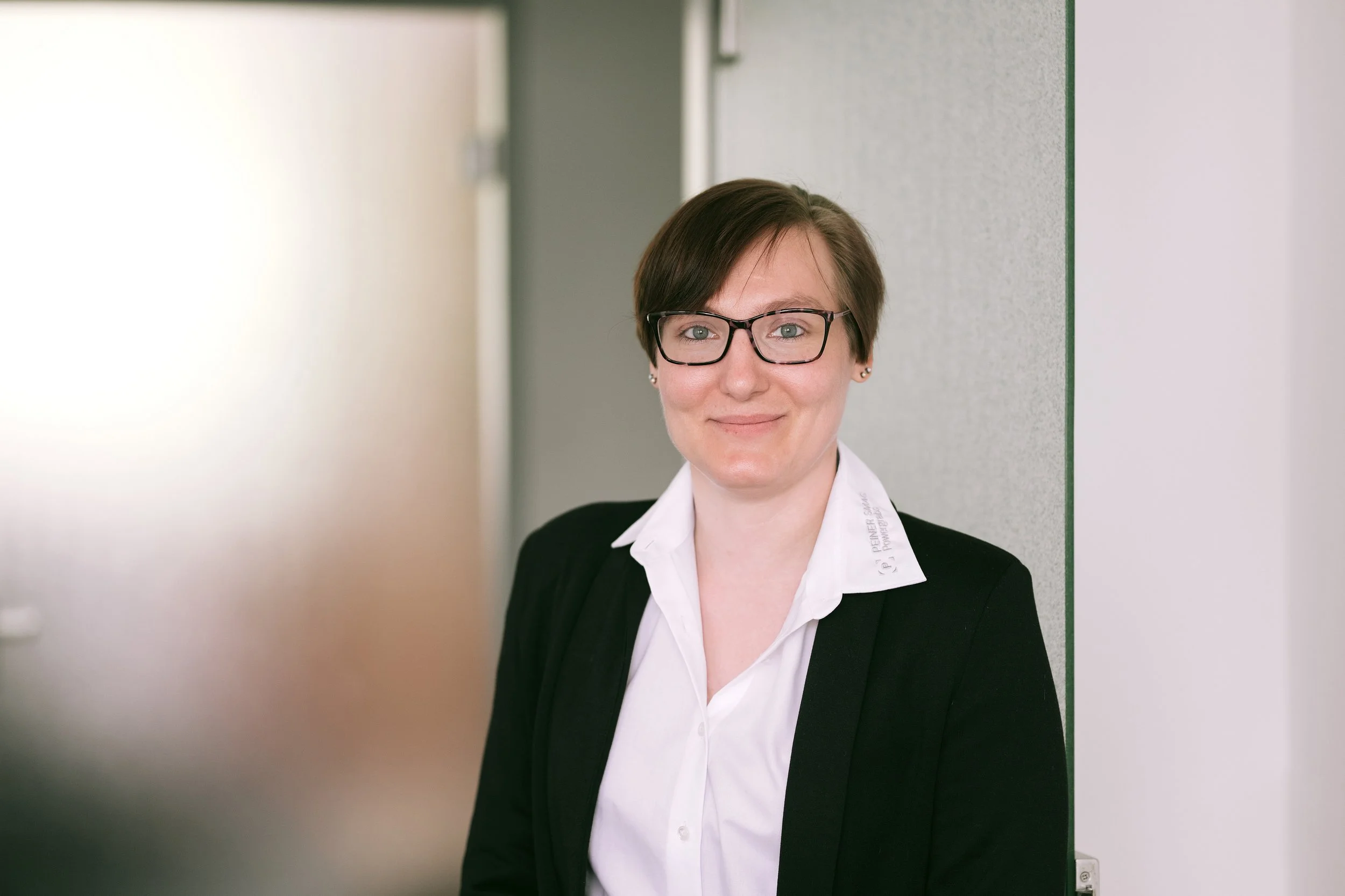 Porträt einer jungen Frau mit Brille, dunklem Kurzhaarschnitt, weißem Hemd und schwarzer Jacke, lächelnd, steht in einem Flur an einer Wand.