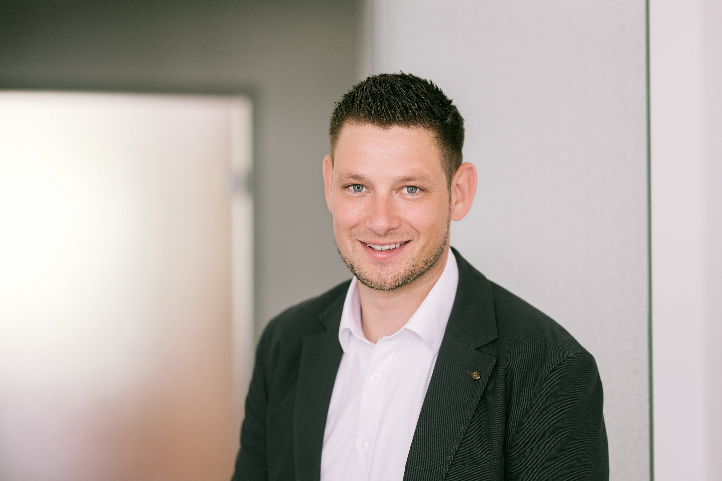 Ein junger Mann mit dunklen, kurzen, gestylten Haaren, trägt einen schwarzen Anzug und ein weißes Hemd, lächelt und steht in einem modernen, hellen Raum.