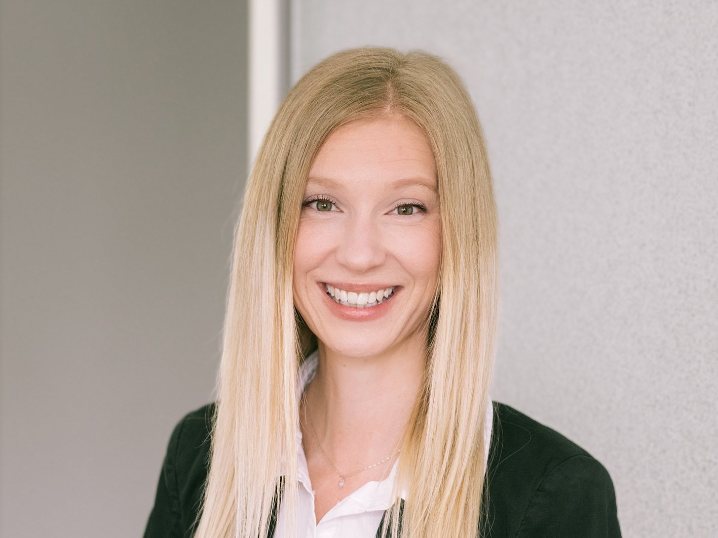 Porträt einer lächelnden Frau mit langem blondem Haar, die eine weiße Bluse und eine schwarze Jacke trägt, vor neutralem Hintergrund.