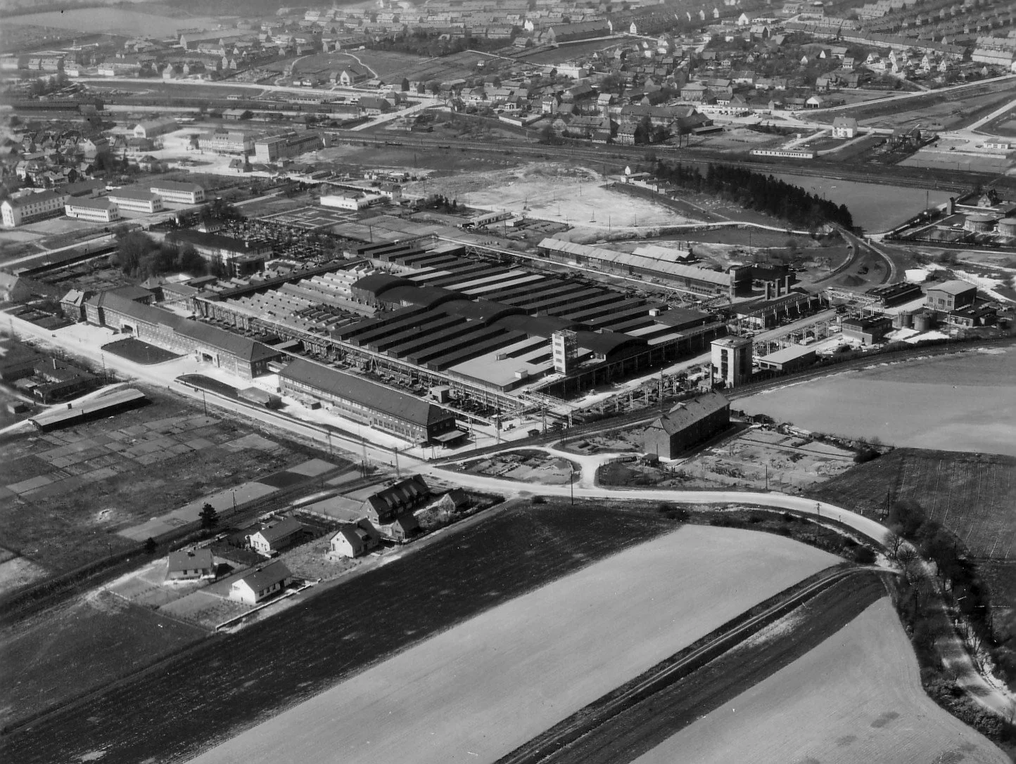 Luftaufnahme einer industriellen Anlage mit mehreren großen Lagerhallen, umgeben von Feldern und Wohnhäusern im Hintergrund.