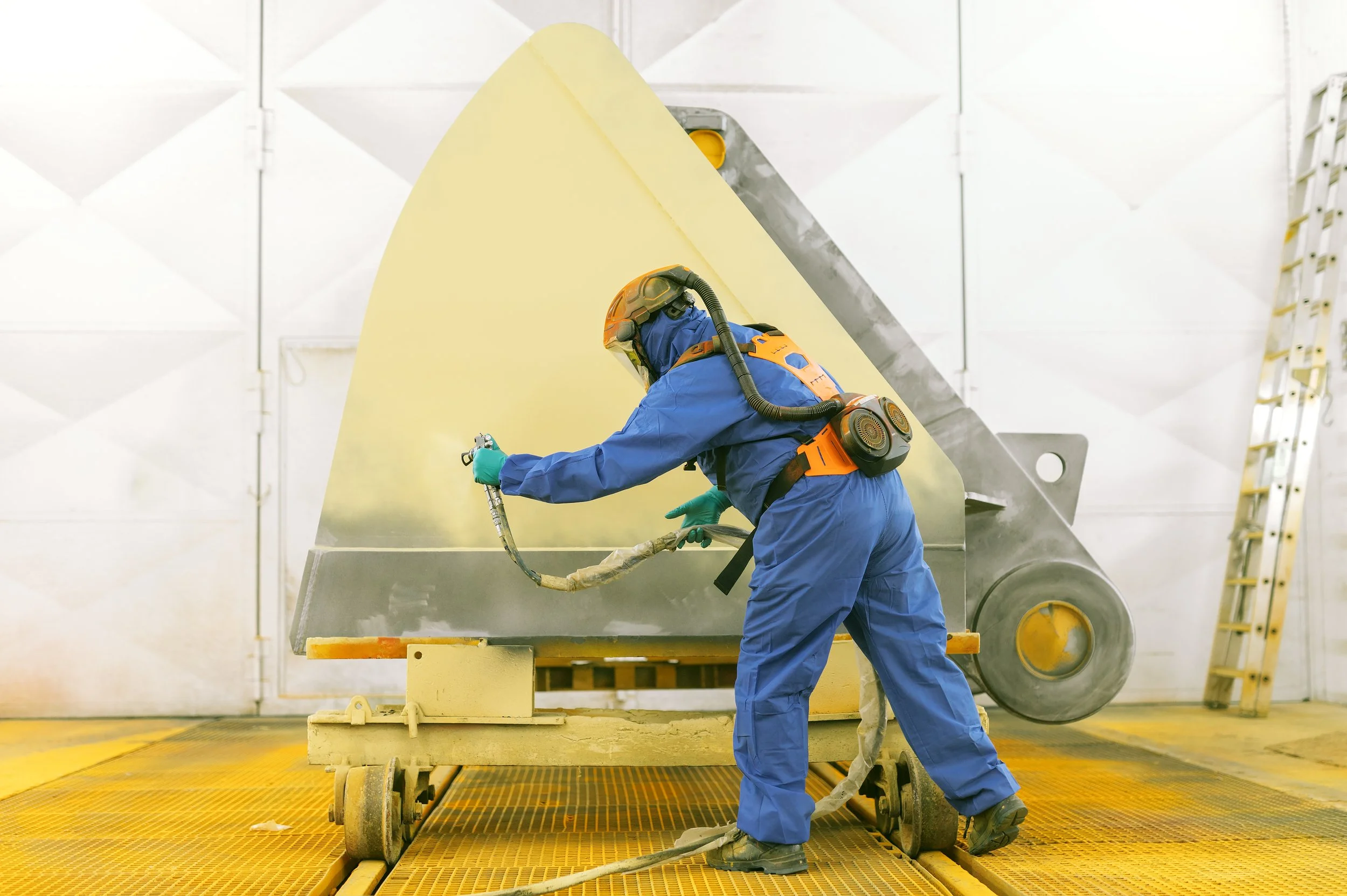 Ein Techniker in blauer Schutzkleidung und Maskierung arbeitet in einer industriellen Anlage mit großen Metallteilen und Heizungen, umgeben von Weißwandpaneelen und einer Leiter im Hintergrund.