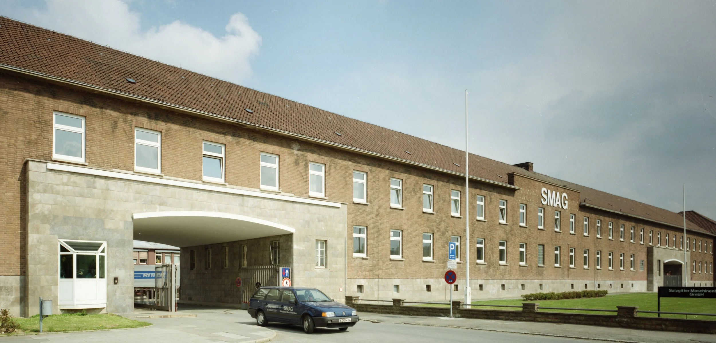 Ein großes, zweistöckiges Firmengebäude aus Backstein mit mehreren Fenstern und einem roten Ziegeldach. Es trägt das Logo 'SMAG' an der Seite. Es gibt einen Durchgang unter einem Bogen aus Stein, einen Parkplatz mit einem schwarzen Firmenwagen und einem Parkverbotsschild.