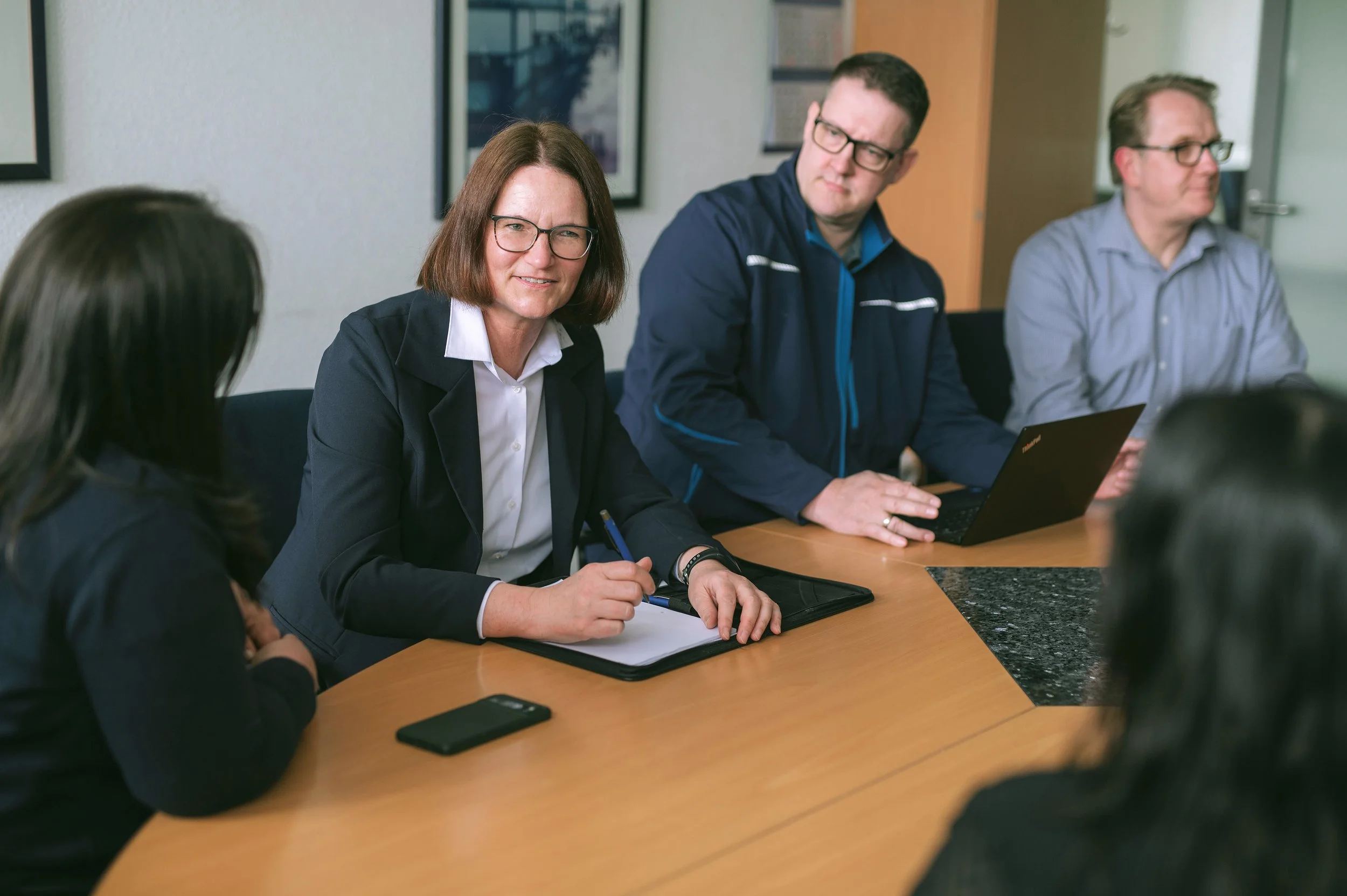 Eine Gruppe von Menschen sitzt an einem Tisch in einem Besprechungsraum, darunter eine Frau mit Brille, die Gesprächspartner zuhört, ein Mann daneben bei einem Laptop, sowie weitere Personen im Hintergrund.