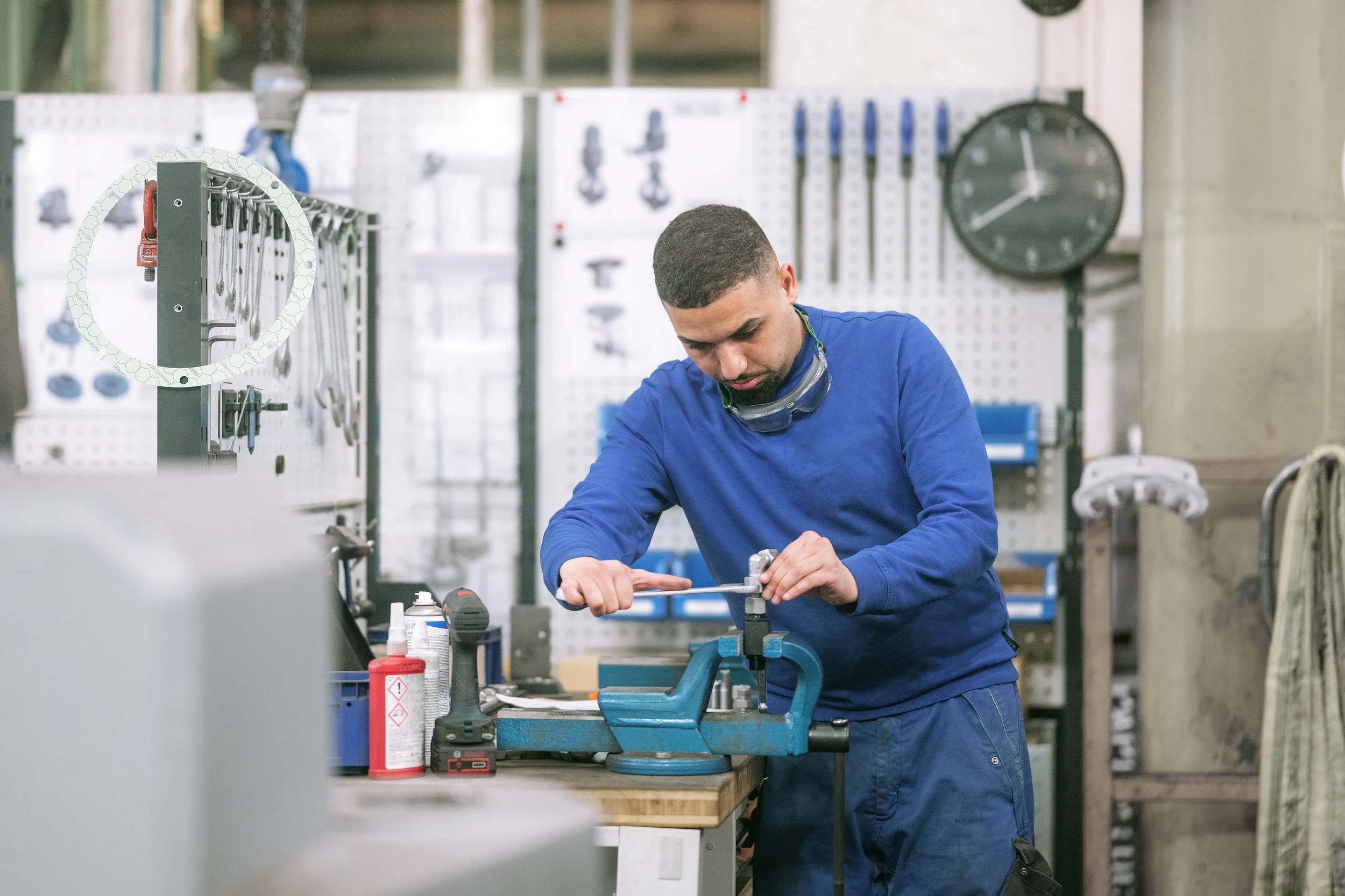 Ein junger Mann arbeitet in einer Werkstatt an einem metallischen Projekt. Er trägt eine blaue Arbeitsjacke und Schutzbrille um den Hals. Im Hintergrund sind Werkzeuge an einer Wand befestigt, es gibt eine große Uhr an der Wand.