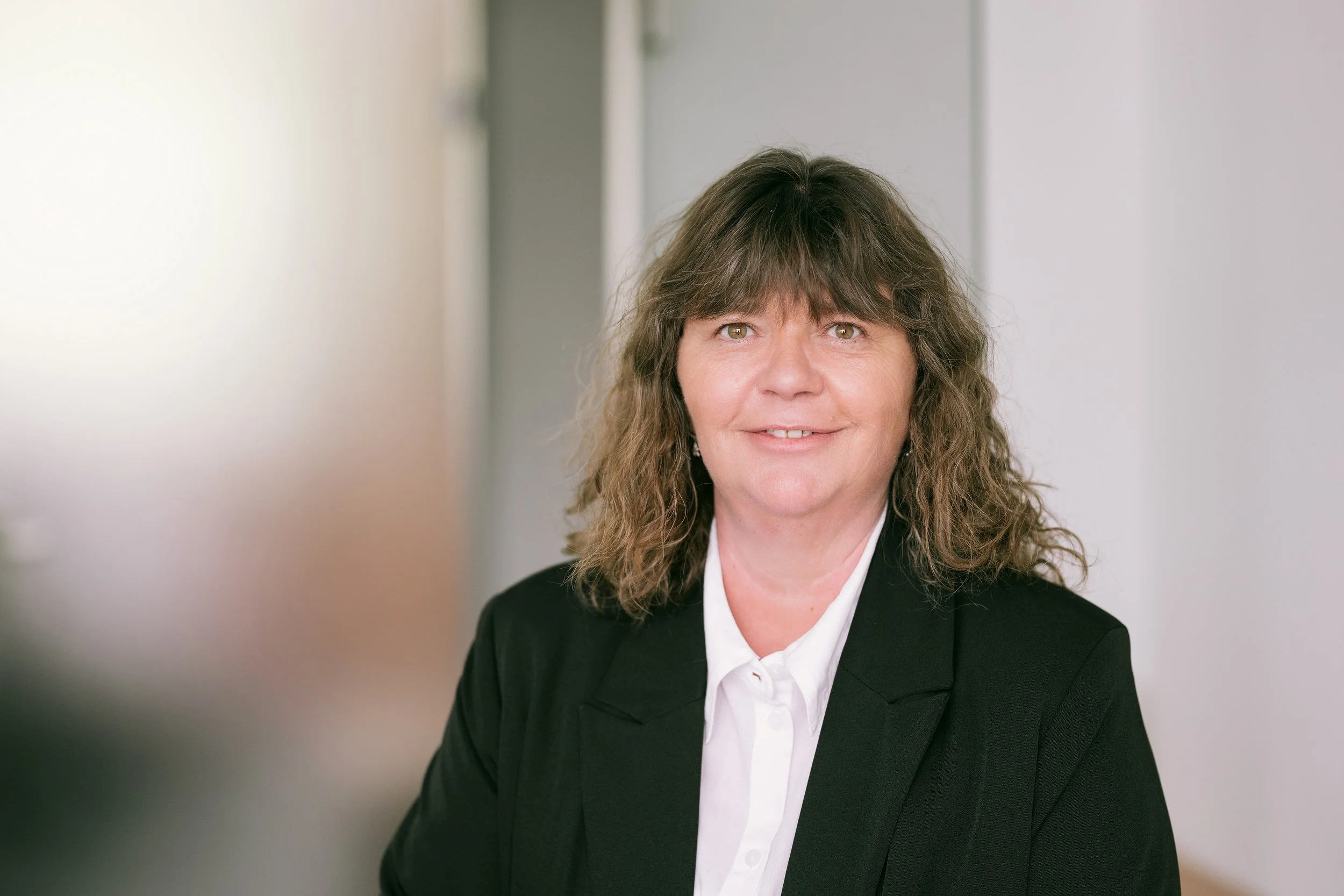 Porträt einer Frau mit braunen, gelockten Haaren, die eine schwarze Jacke und weiße Bluse trägt, vor einem neutralen Hintergrund.