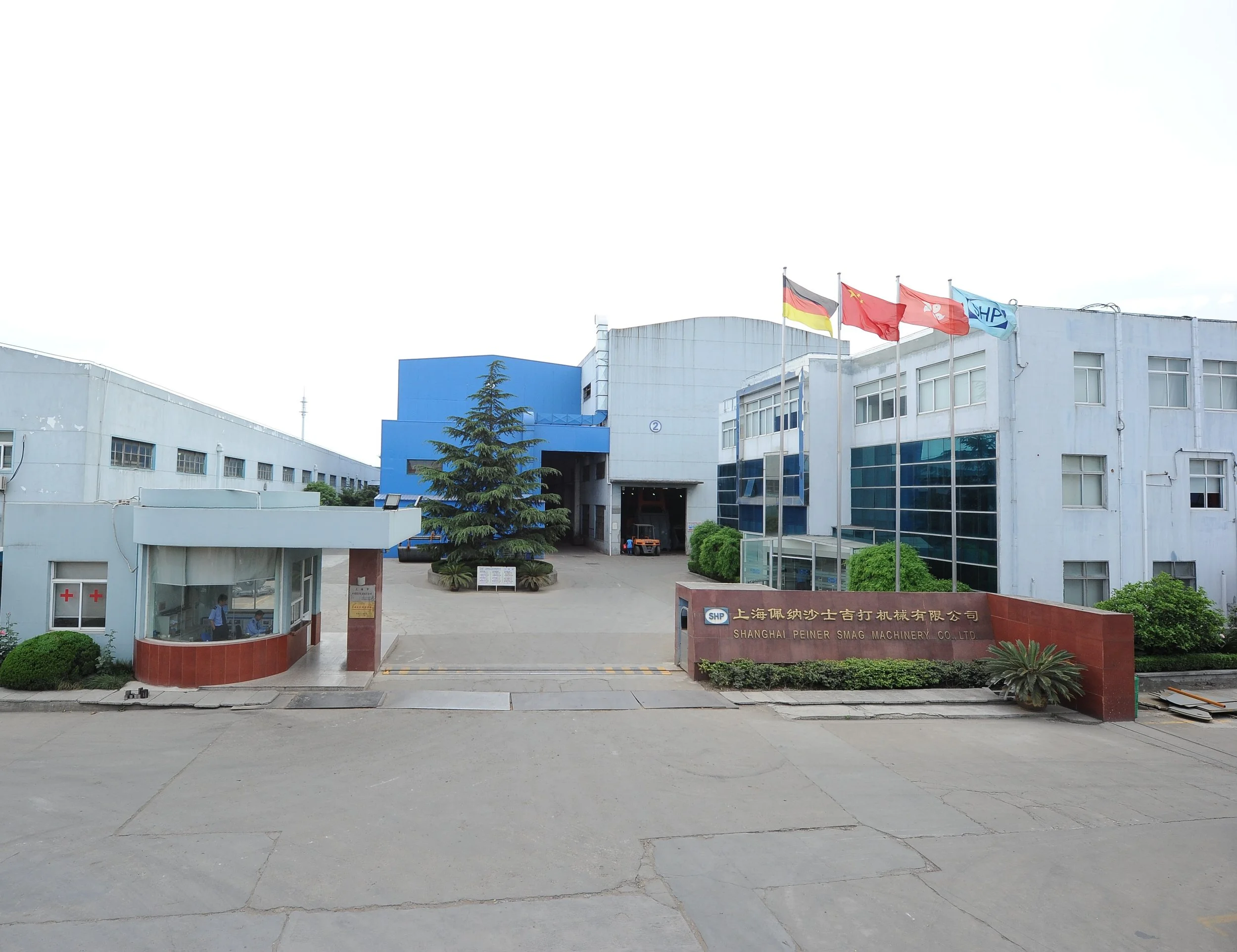 Das Bild zeigt das Gebäude der Shanghai Peiner Smag Machinery Co., Ltd., mit mehreren Fahnen, darunter die deutsche, chinesische und eine blaue Fahne. Vor dem Gebäude sind Büros und ein Eingang, umgeben von einigen Büschen und einem kleinen Baum.