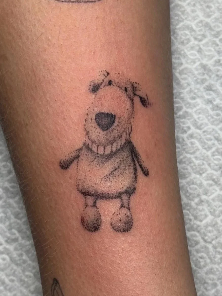 Tattoo van een eenvoudig gestileerd hondje met een grote neus, dat een brede glimlach heeft en een jurk draagt.