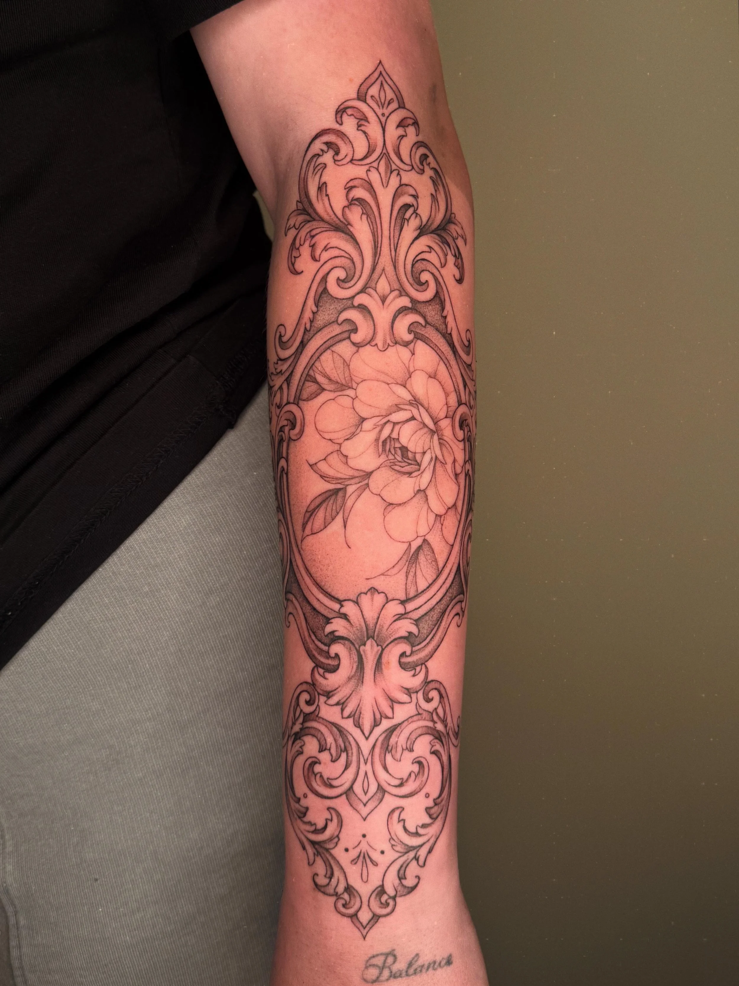 Tatoeage op de onderarm met sierlijke patronen en een bloem, en het woord 'Balans' onder de bloem.