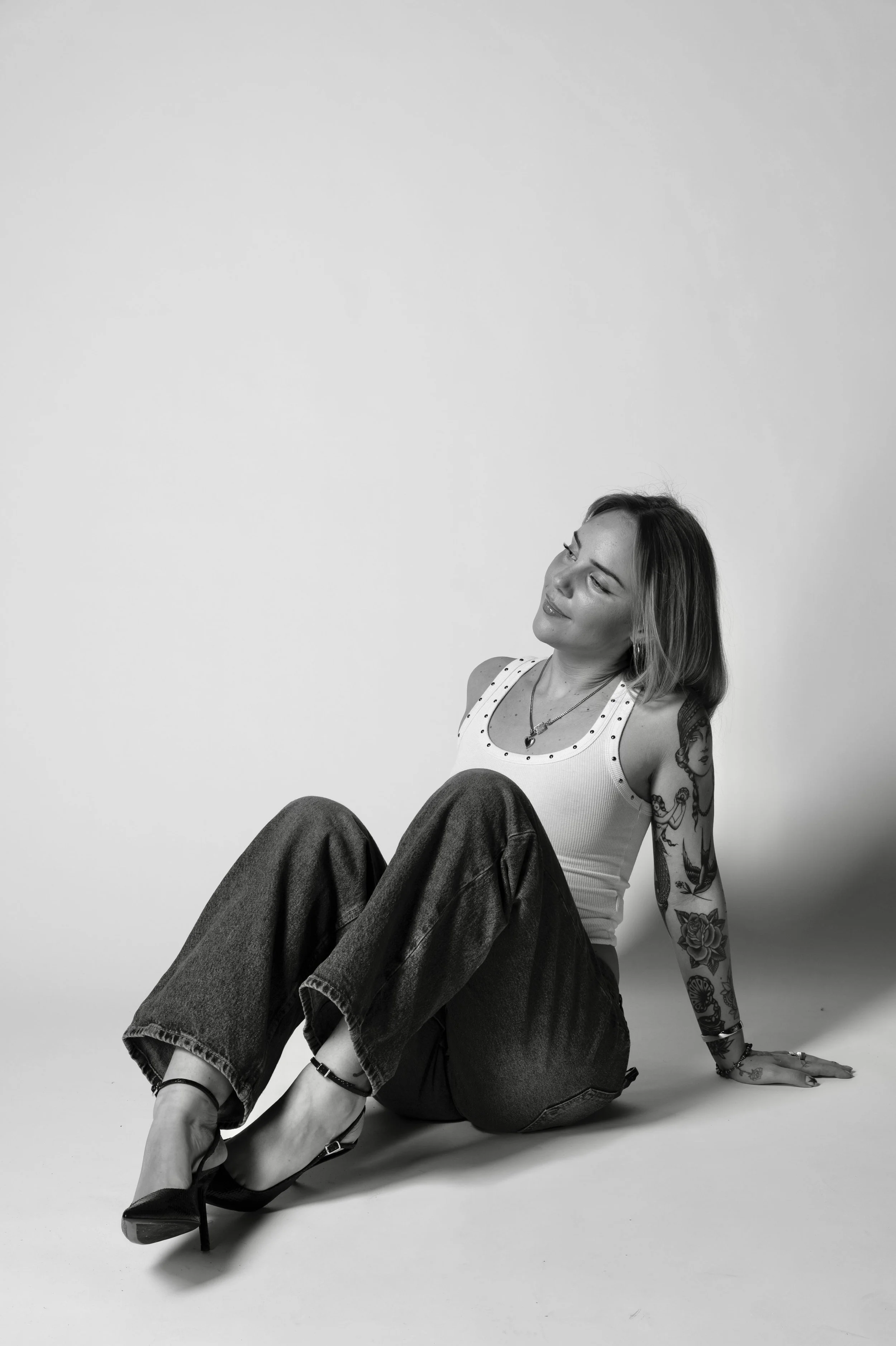 Zwarte en witte foto van een vrouw met tattoos, zittend op de grond, lachend en gekleed in een witte top, loszittende jeans en hoge hakken.