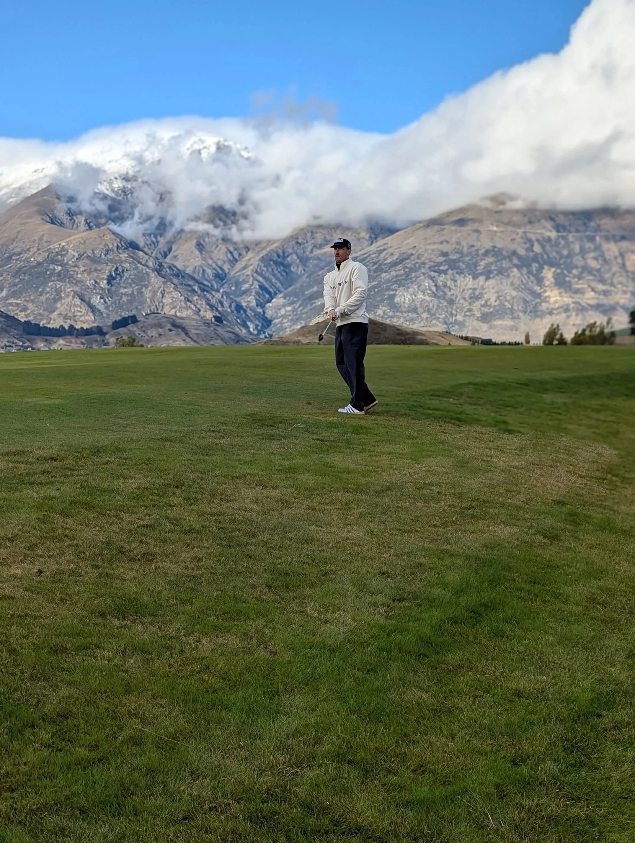 A man in a white jacket and dark pants holding a golf club on a grassy golf course in Queenstown, New Zealand with mountains and clouds in the background.