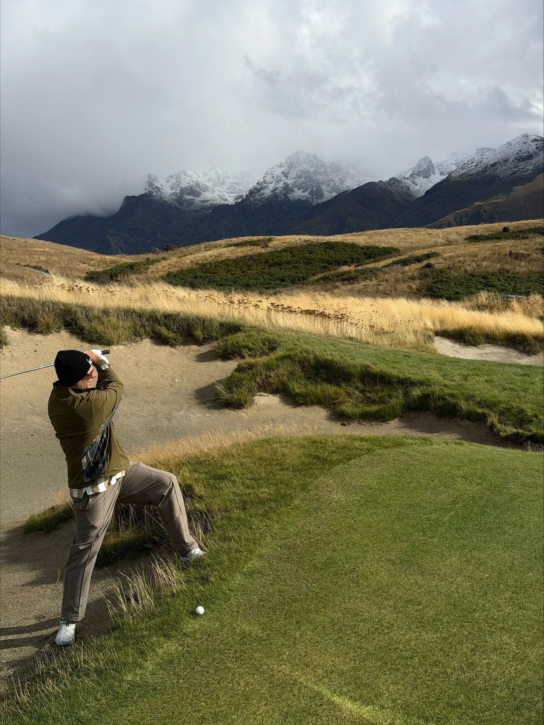 A man playing golf in Queenstown, New Zealand on a course with green grass and sand traps, with mountains and cloudy sky in the background.