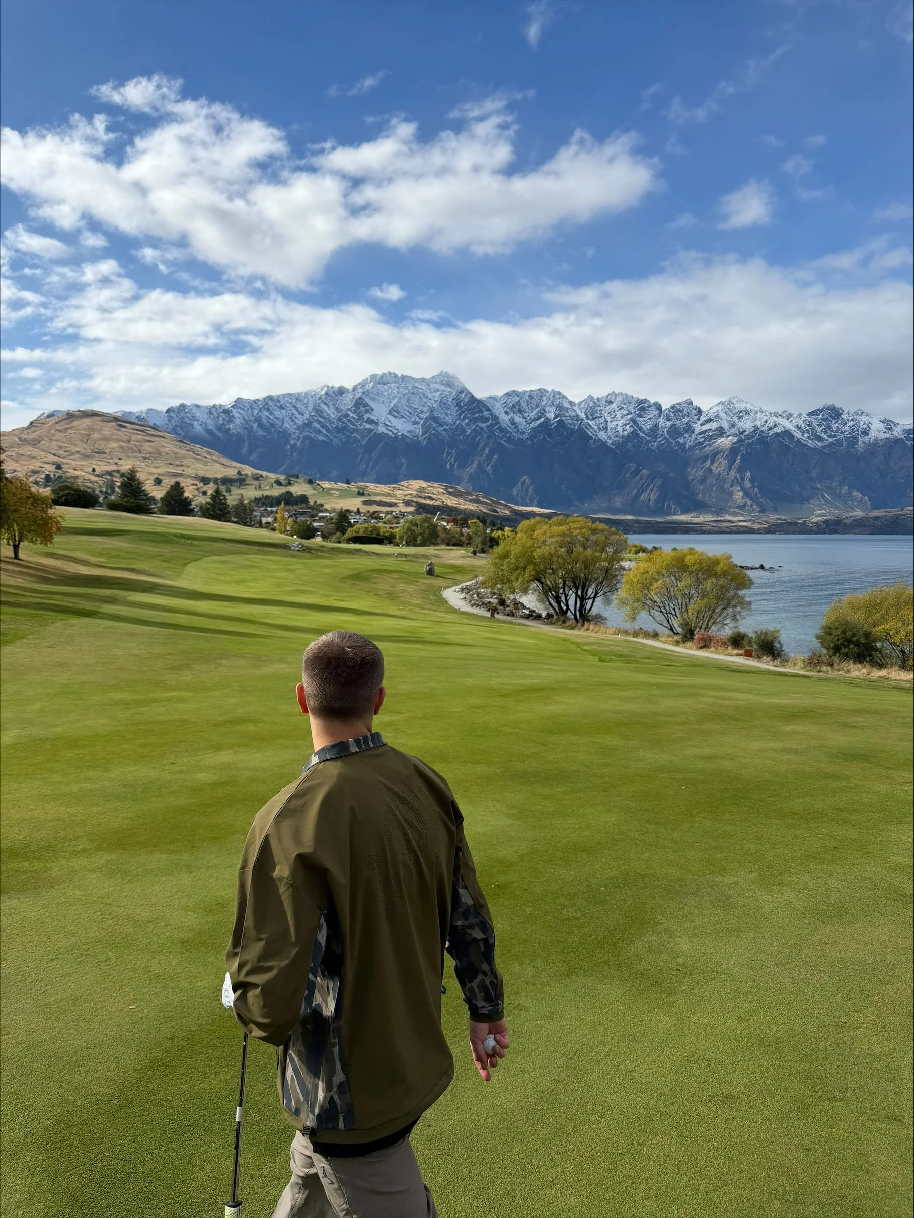 A man on a Queenstown, New Zealand golf course holding a golf ball and club, with mountains and a lake in the background, under a partly cloudy sky.