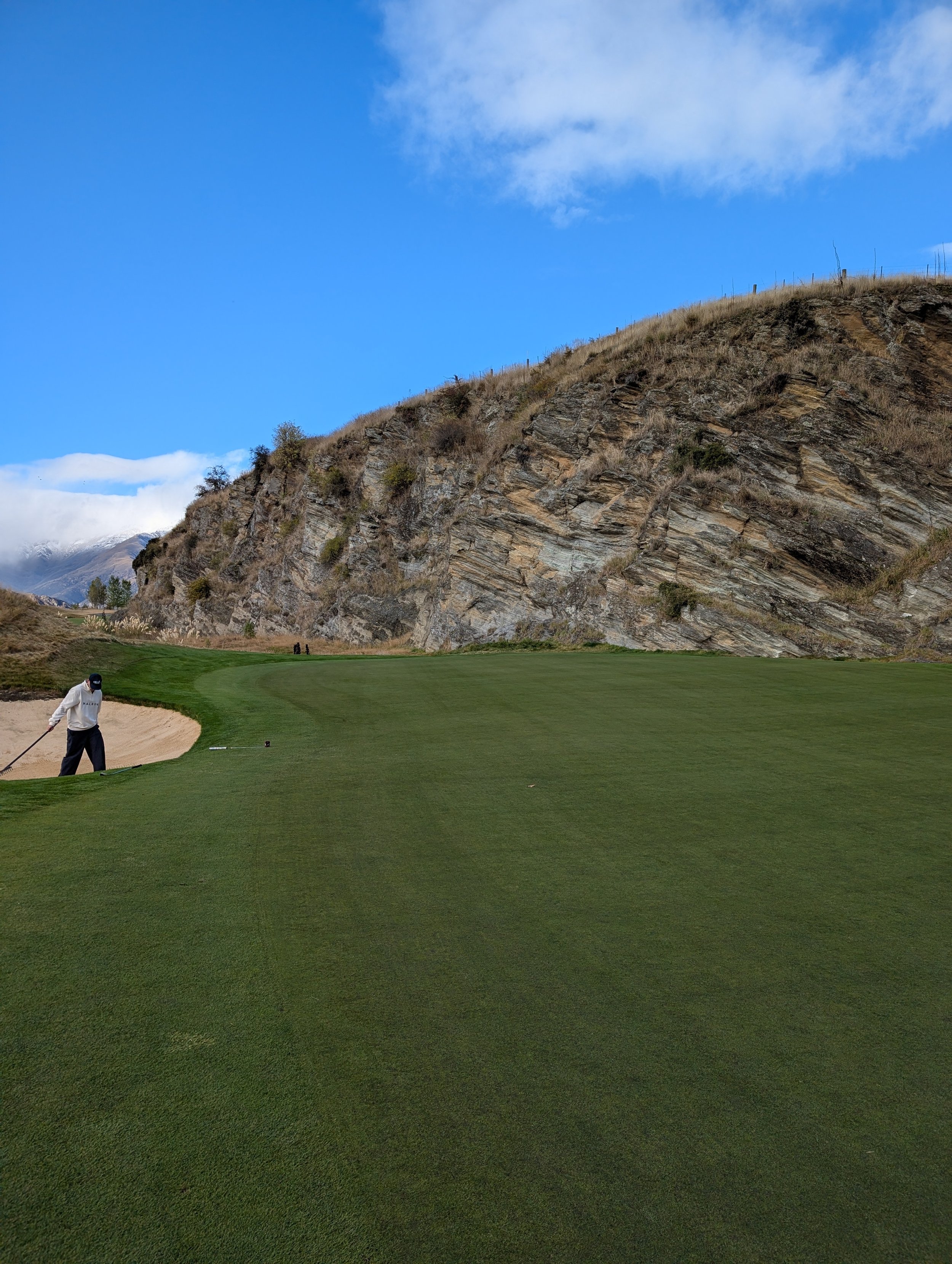 A golfer preparing to hit a shot on a golf course in Queenstown, New Zealand with a grassy fairway, rock formations, and mountains in the background under a partly cloudy sky.