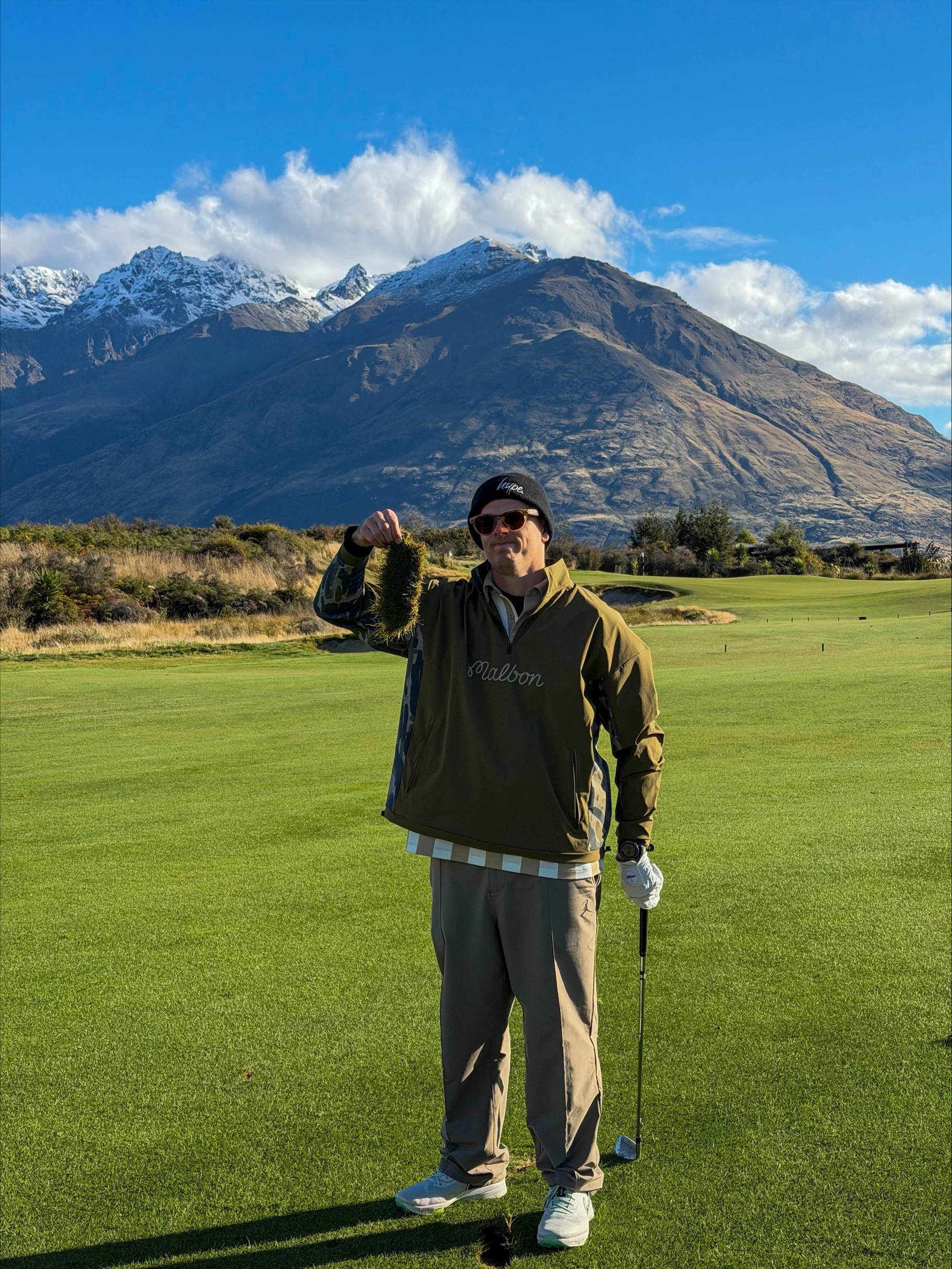 A man standing on a golf course in Queenstown, New Zealand holding a golf club and a piece of his grass, with a mountain range in the background under a mostly blue sky with some clouds.