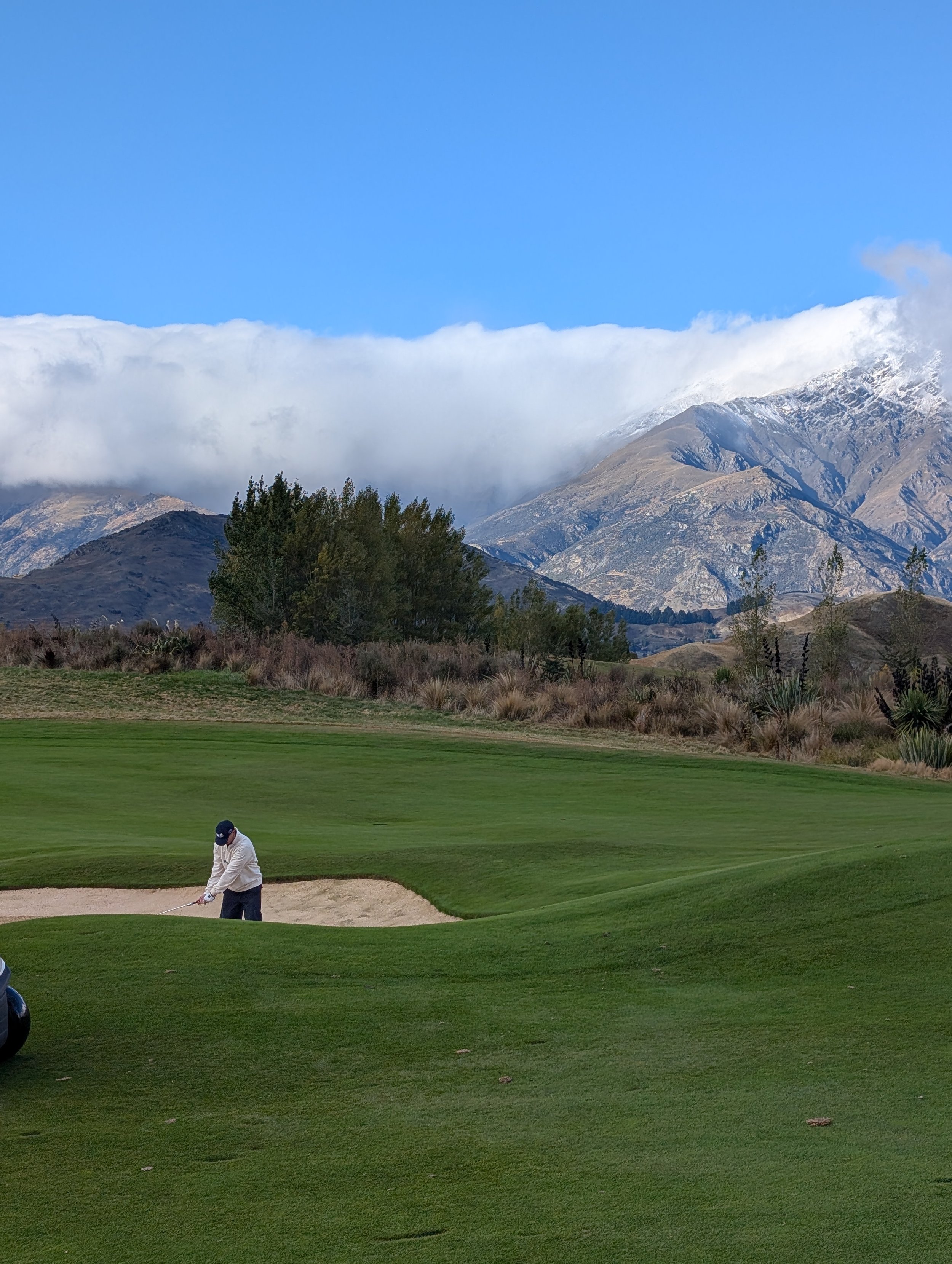 A golfer in white attire and a cap playing golf on a well-maintained course with lush green grass, greenery, and mountains in the background, under a clear blue sky with some clouds.
