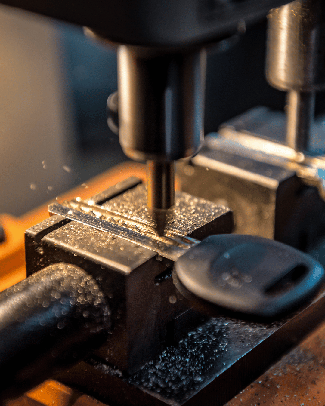 Close-up of a drill making a hole in a metal piece mounted on a vice, with metal shavings scattered around.