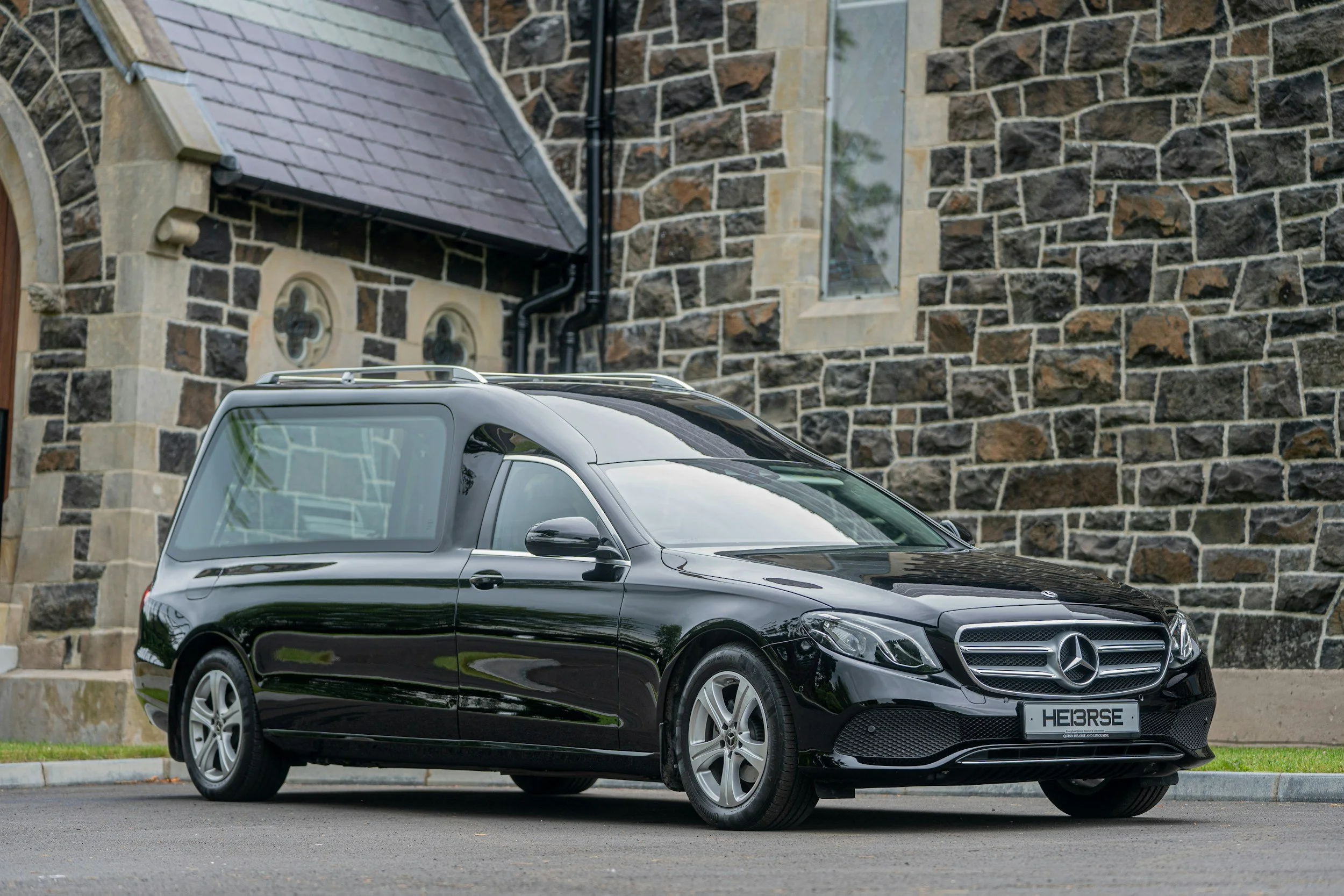Black Mercedes-Benz hearse parked in front of a building with stone walls and small, arched windows.