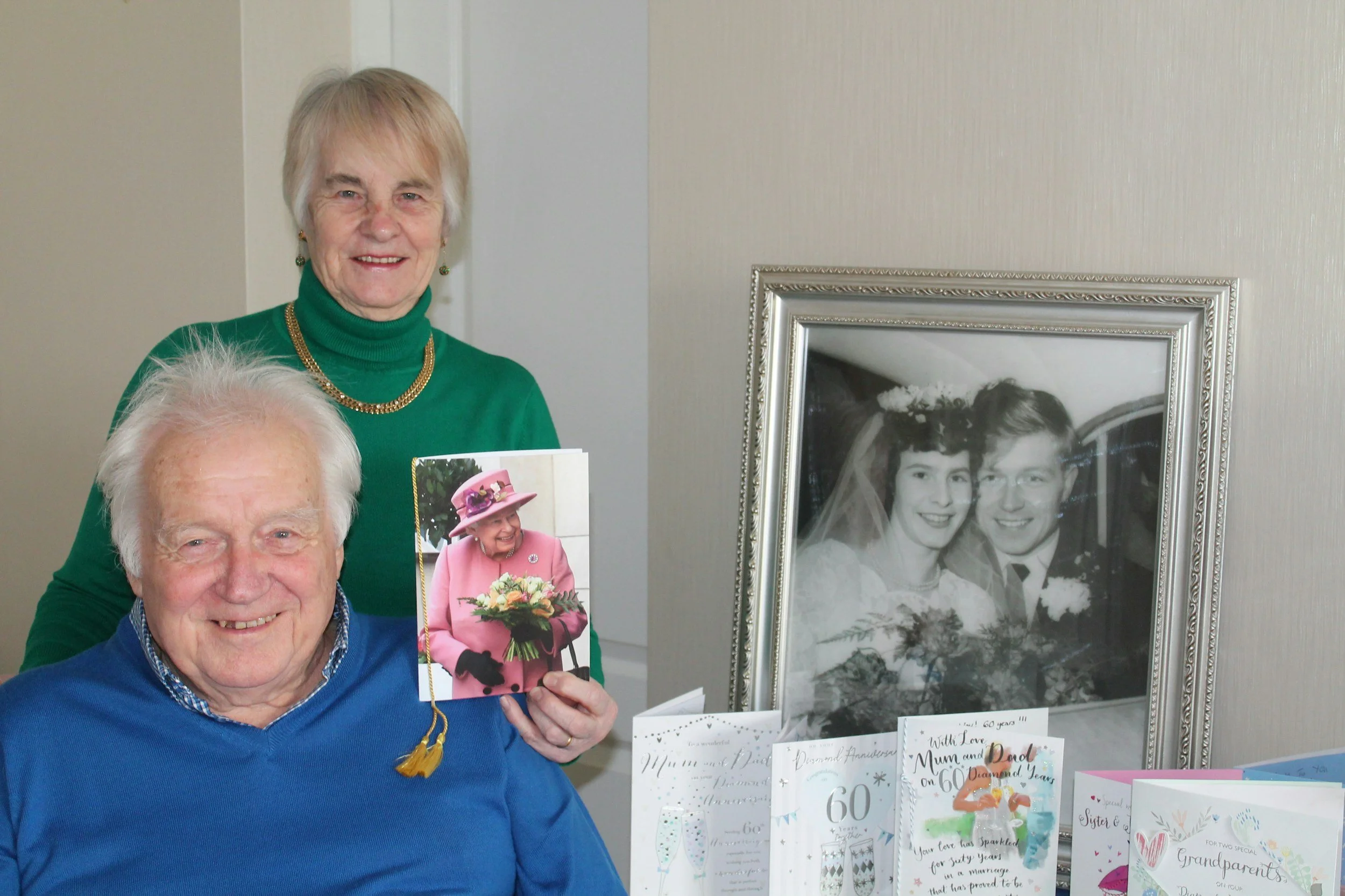 An elderly man and a woman at a celebration, holding greeting cards and photographs, with a display of anniversary cards and a framed black-and-white wedding photo on the wall behind them.
