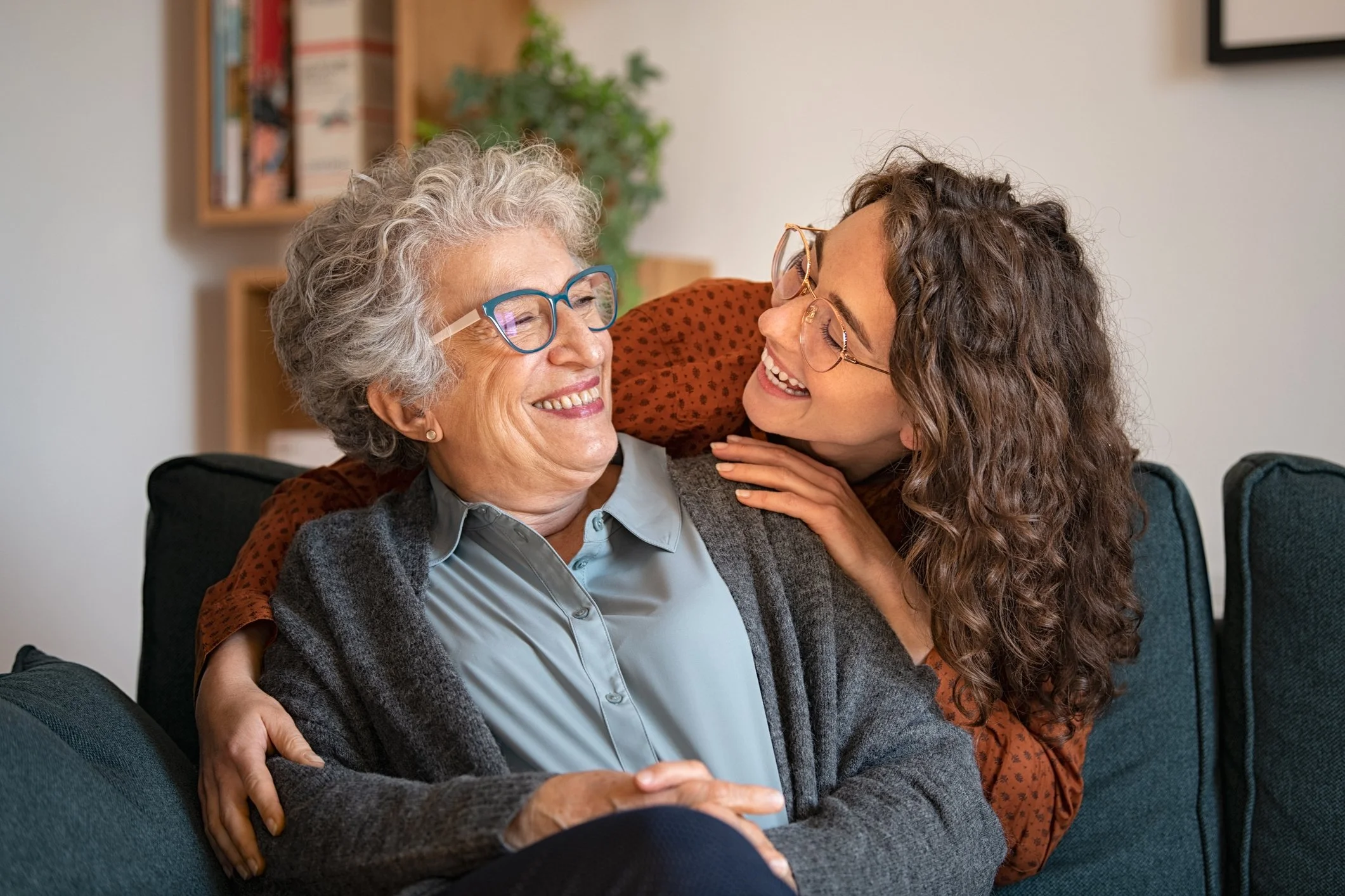An elderly woman with gray, curly hair and glasses smiling as she talks with a younger woman with long, curly hair and glasses. They are sitting close together on a couch in a living room, sharing a joyful moment.