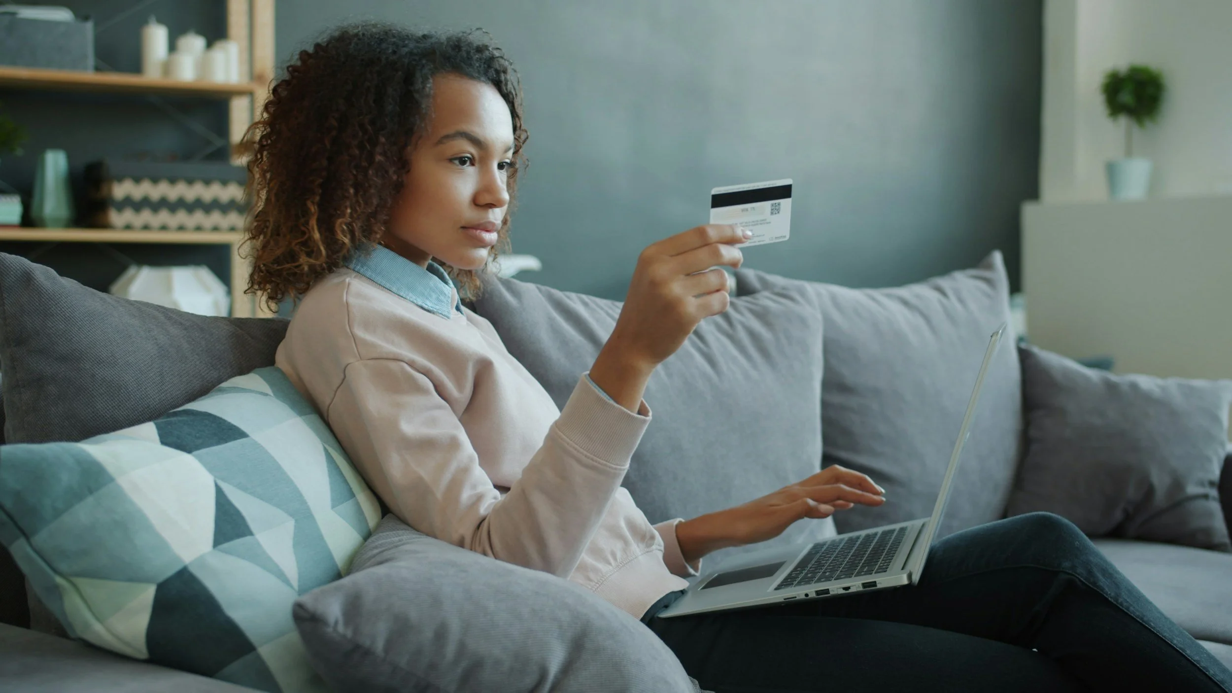 A woman sitting on a gray couch holding a credit card in her right hand while using a laptop on her lap in a living room.