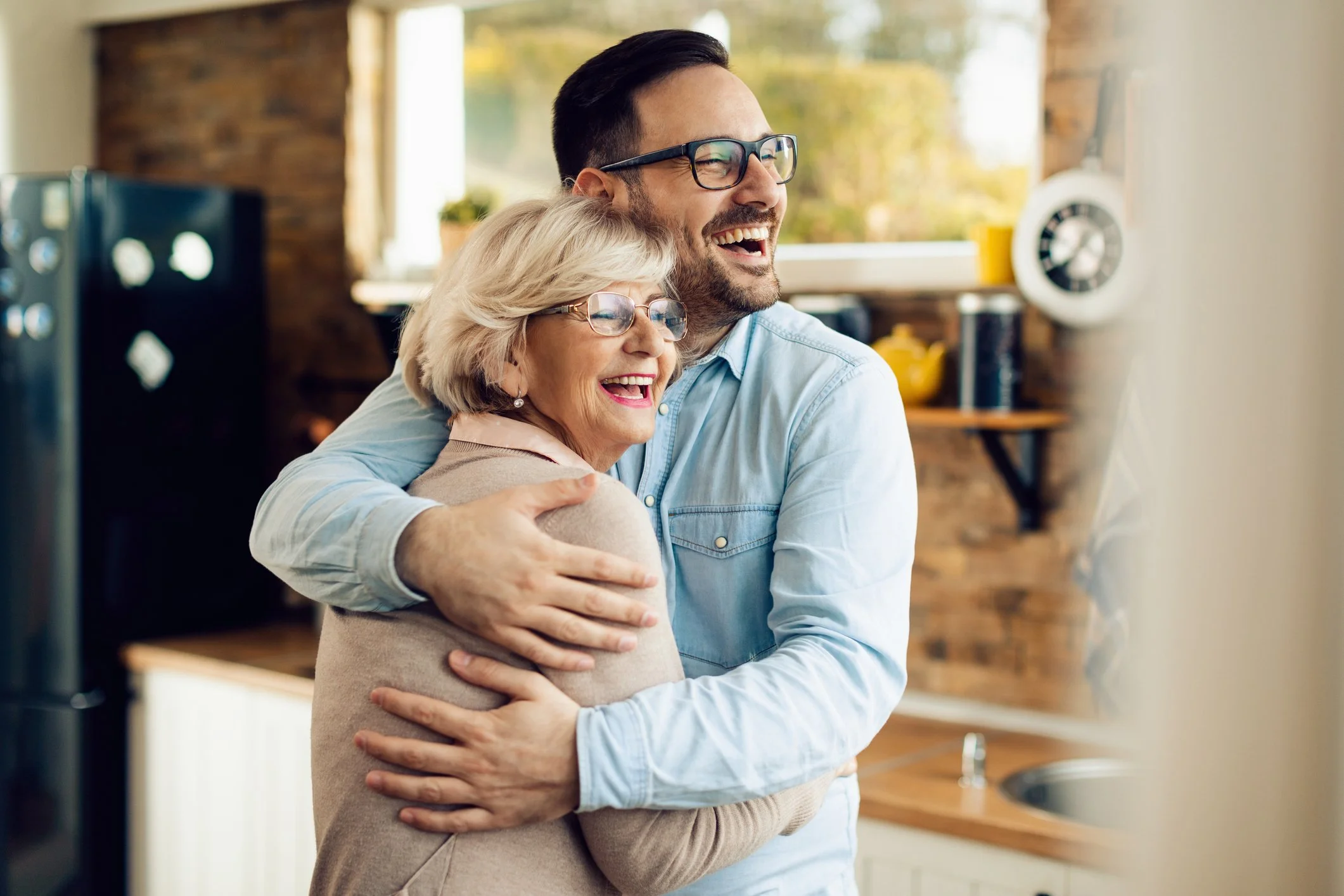 A young man and an elderly woman happily hugging in a kitchen with a window showing trees outside.