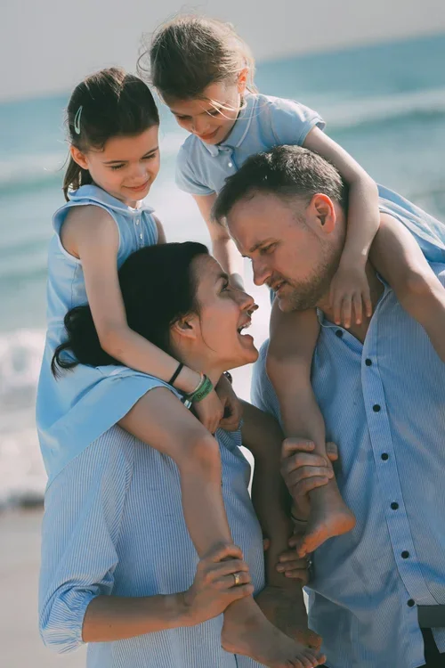 A family enjoying a day at the beach, with two young girls riding on their parents' shoulders and smiling.