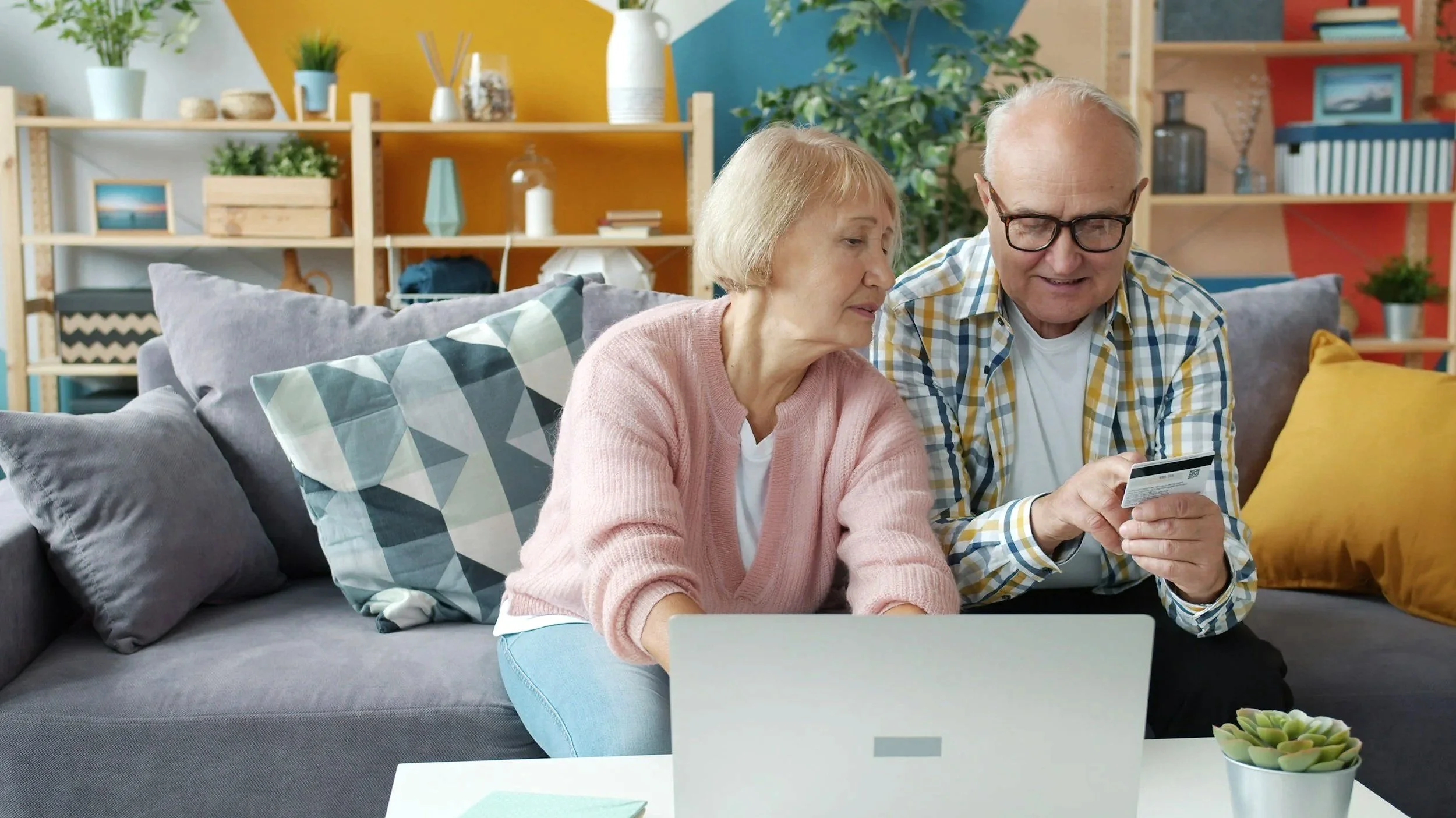An elderly couple sitting on a gray sofa in a living room, looking at a smartphone together.