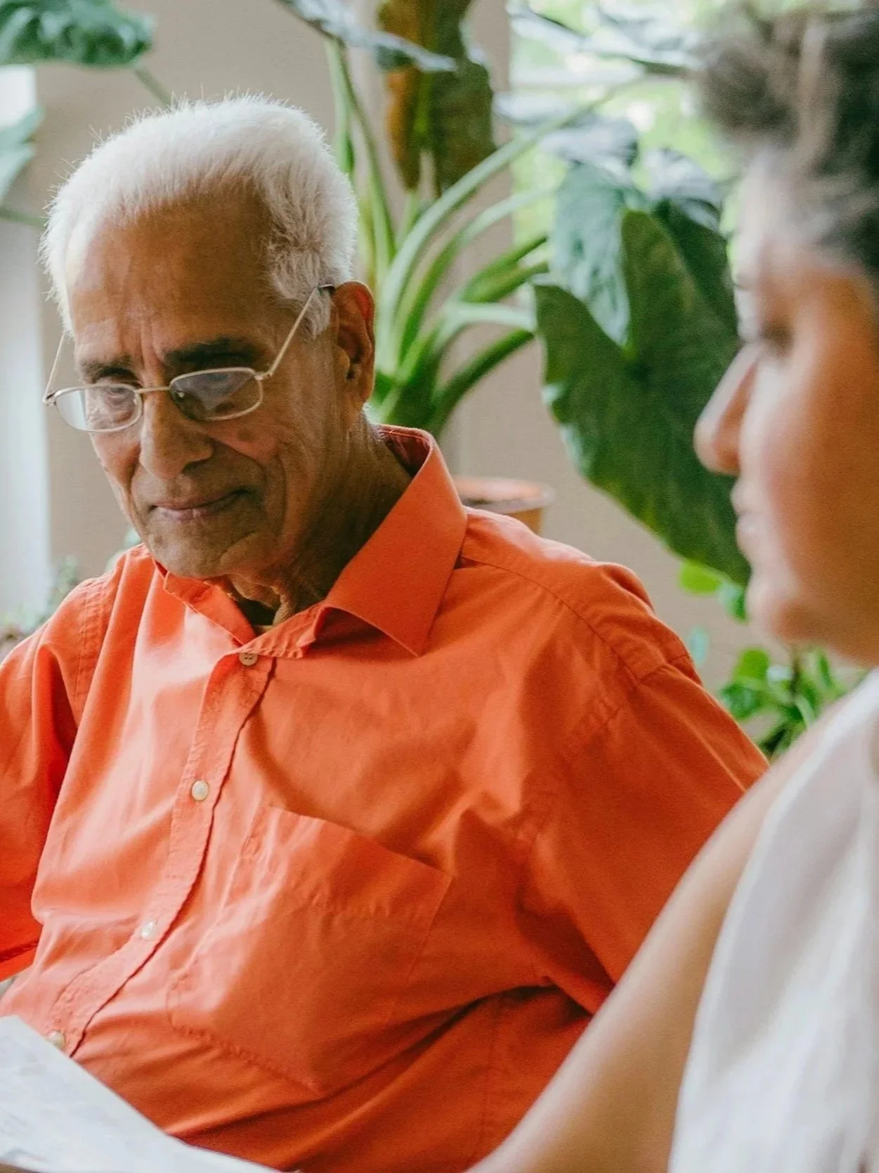 An elderly man with white hair and glasses wearing an orange shirt, seated indoors with green plants in the background.