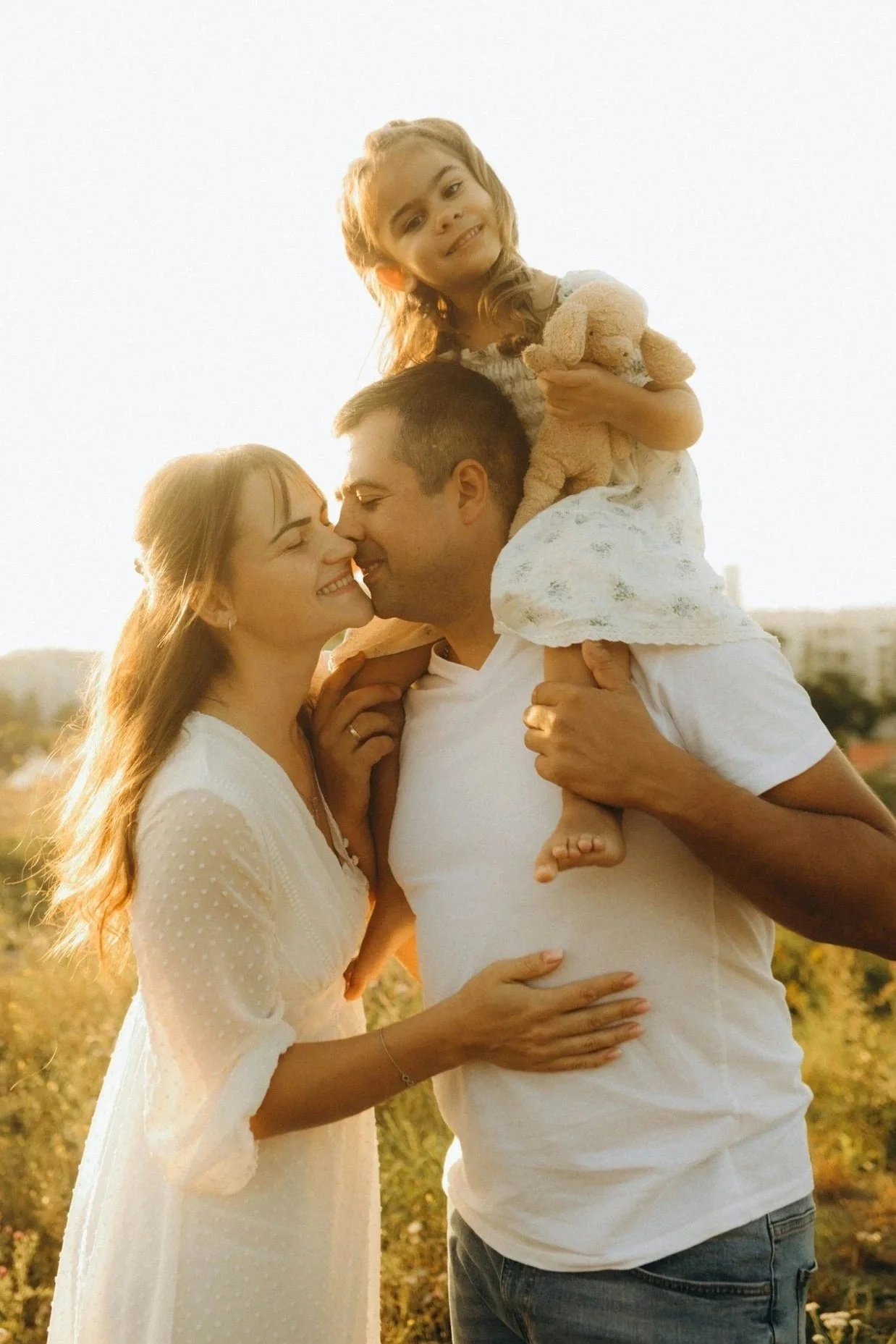 A family of three enjoying a moment outdoors during sunset, with a woman and man kissing and a young girl sitting on the man's shoulders holding a stuffed animal.