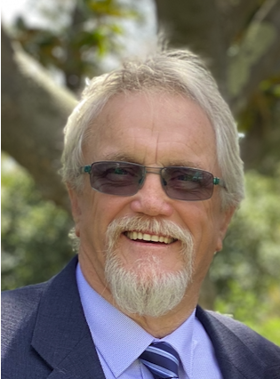 A smiling elderly man with gray hair and a beard, wearing sunglasses, a blue suit, and a striped tie outdoors.