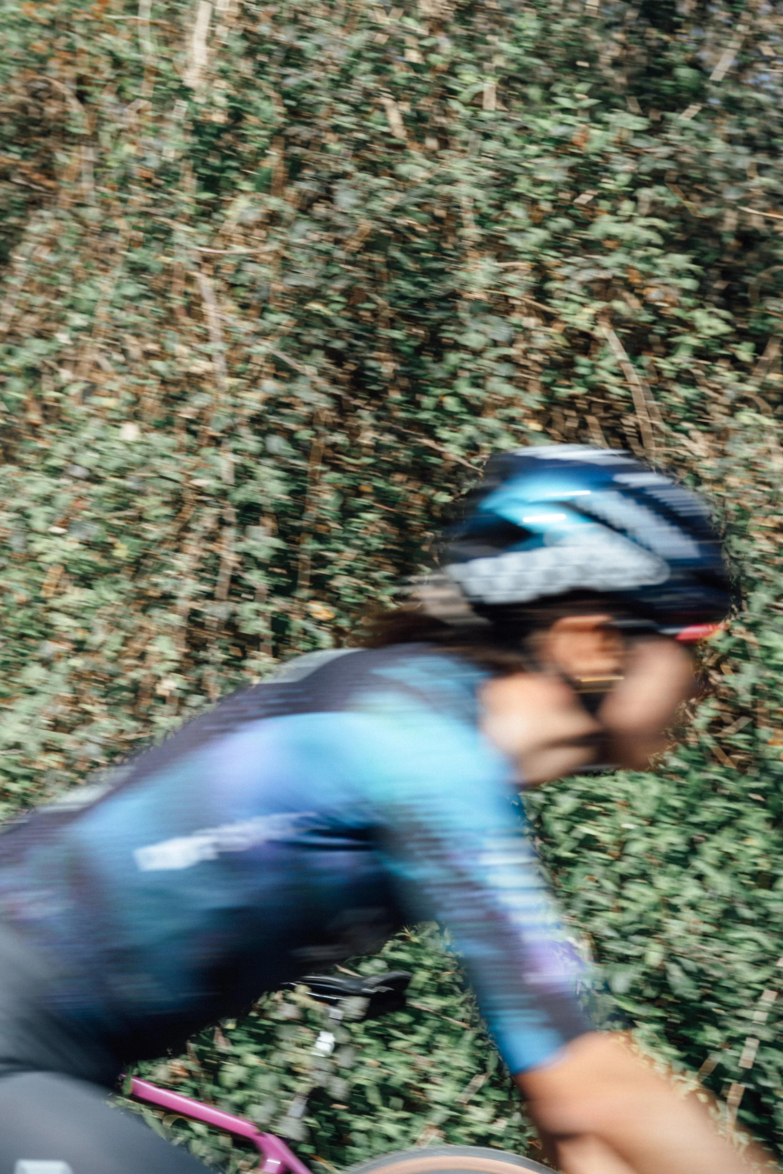 Blurry photo of a person wearing a helmet and riding a bicycle through a wooded area with green foliage.