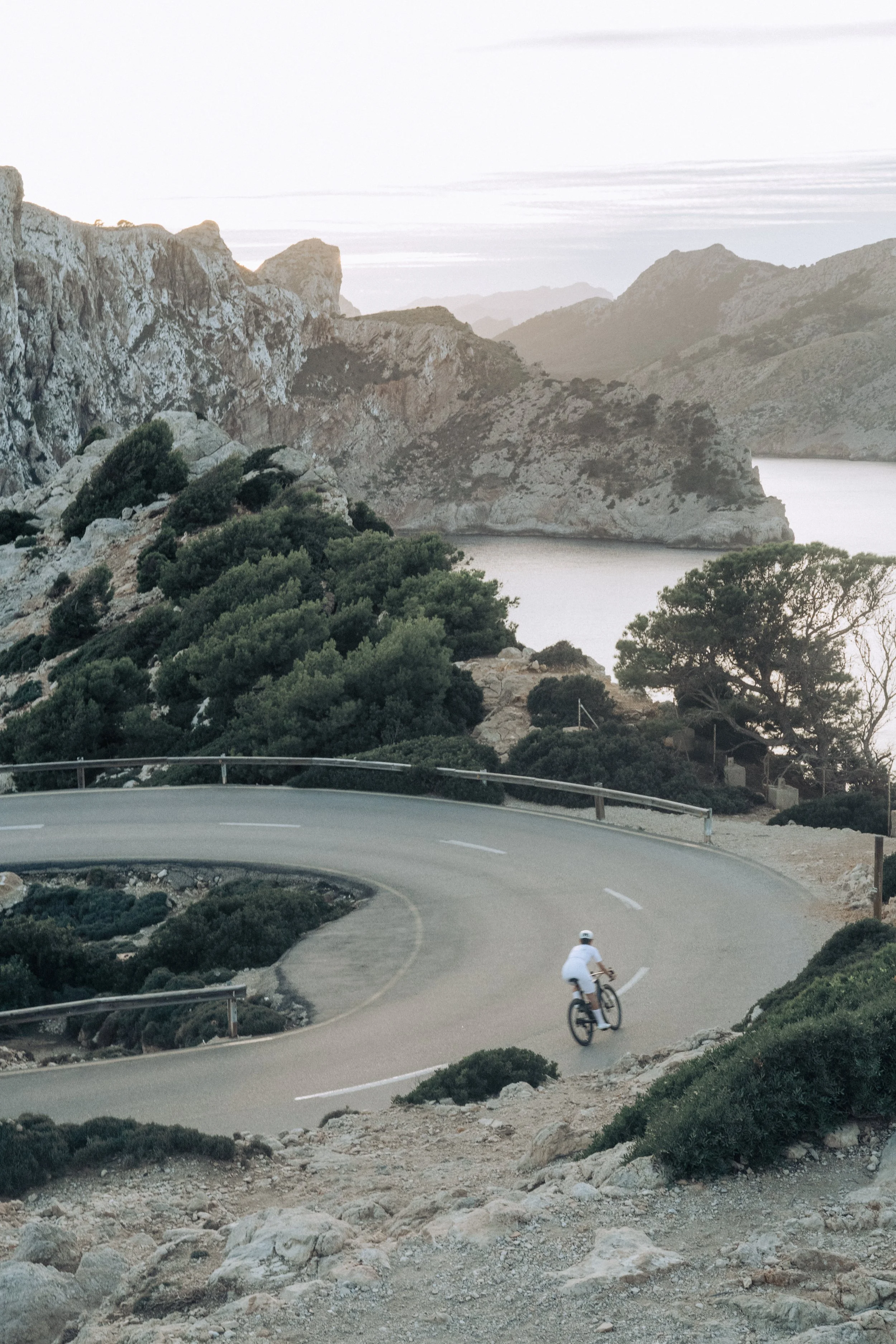 A cyclist riding a bicycle on a winding mountain road with rocky terrain and lush green bushes. In the background, there are rugged mountains and a lake. The scene appears to be during daytime with cloudy weather.