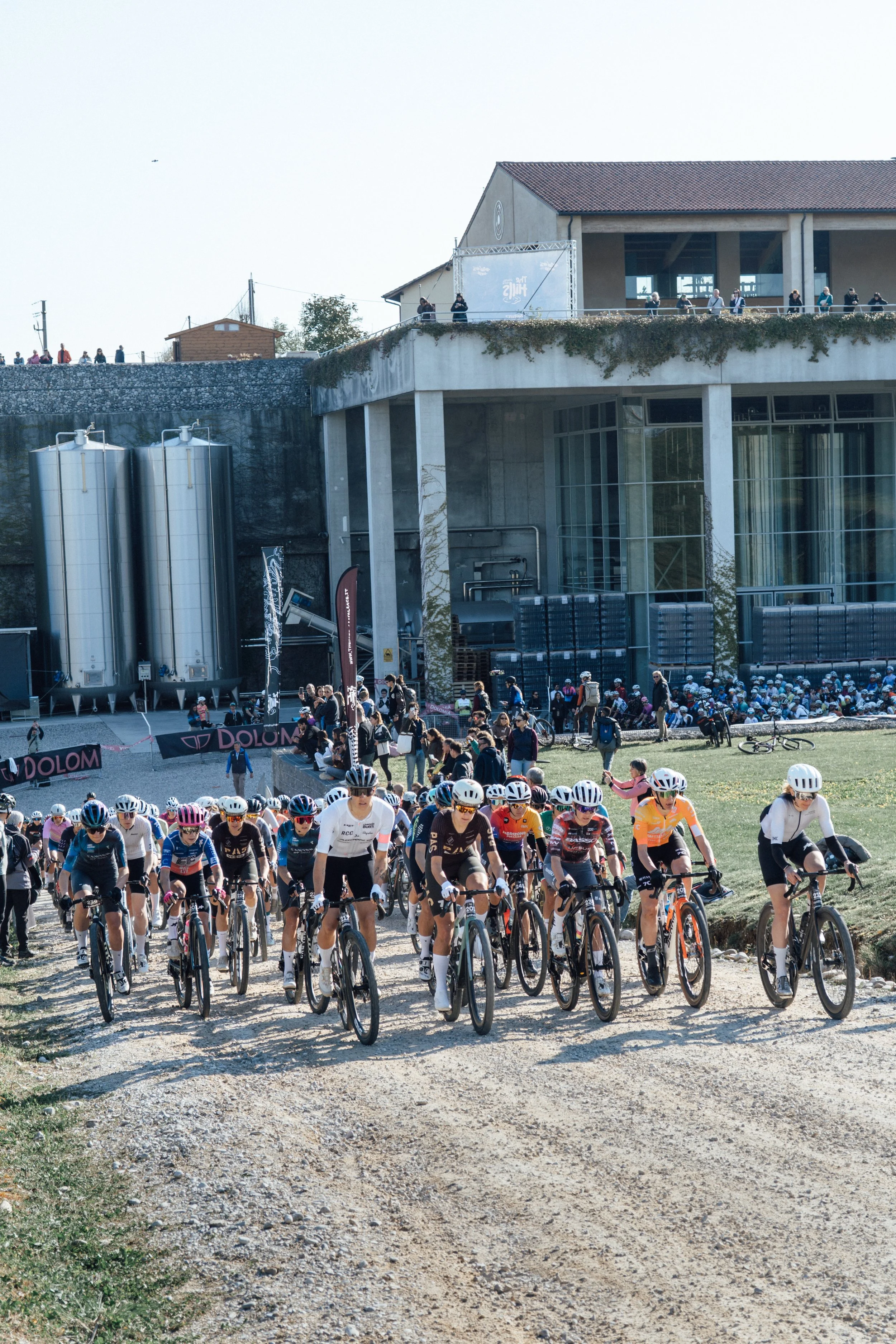 A large group of cyclists participating in a race or event on a dirt path with a building and spectators on an upper balcony in the background.
