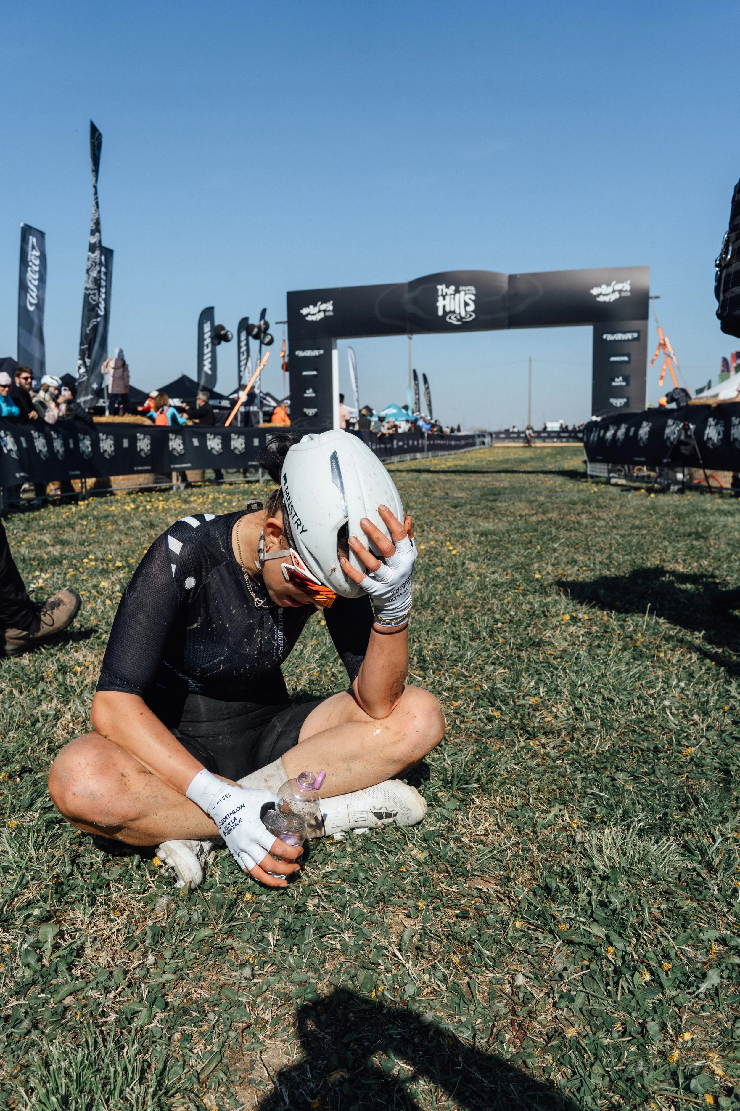 A cyclist, wearing a helmet and athletic gear, sits on the ground with one hand on her head and a water bottle in her other hand at an outdoor cycling event. There are banners, flags, a finish line arch, and spectators in the background under a clear blue sky.
