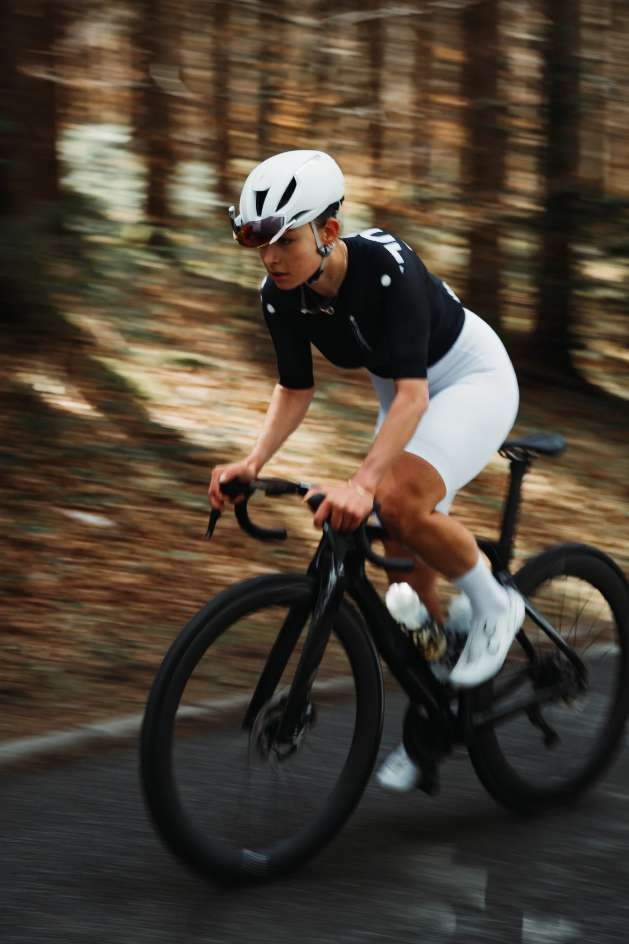 A person riding a bicycle on a paved path through a wooded area with fallen leaves, wearing a white helmet, black cycling jersey, white shorts, and white shoes, with a motion blur indicating speed.