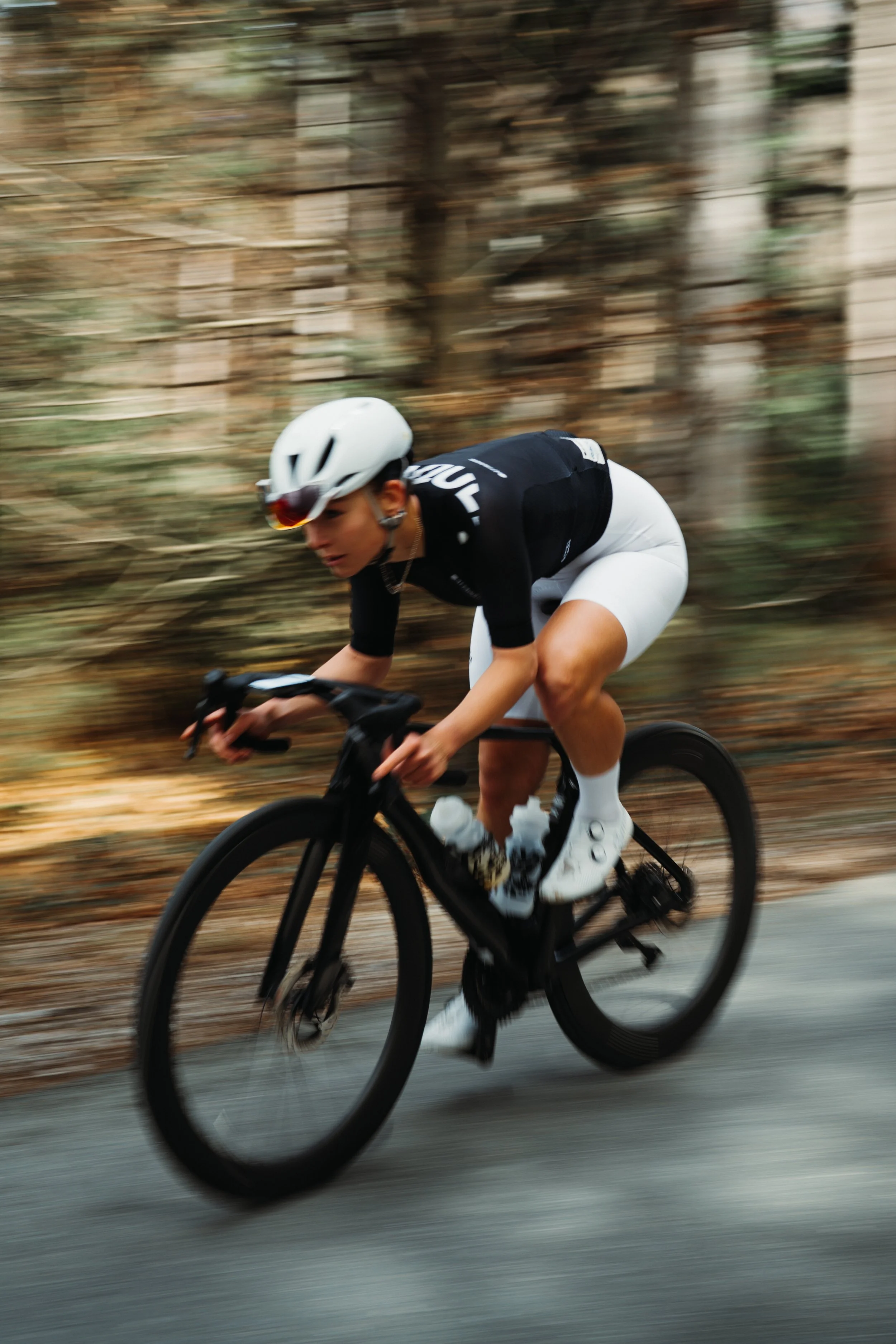 A cyclist wearing a black and white outfit with a white helmet riding a black road bike through a wooded area with autumn leaves falling.