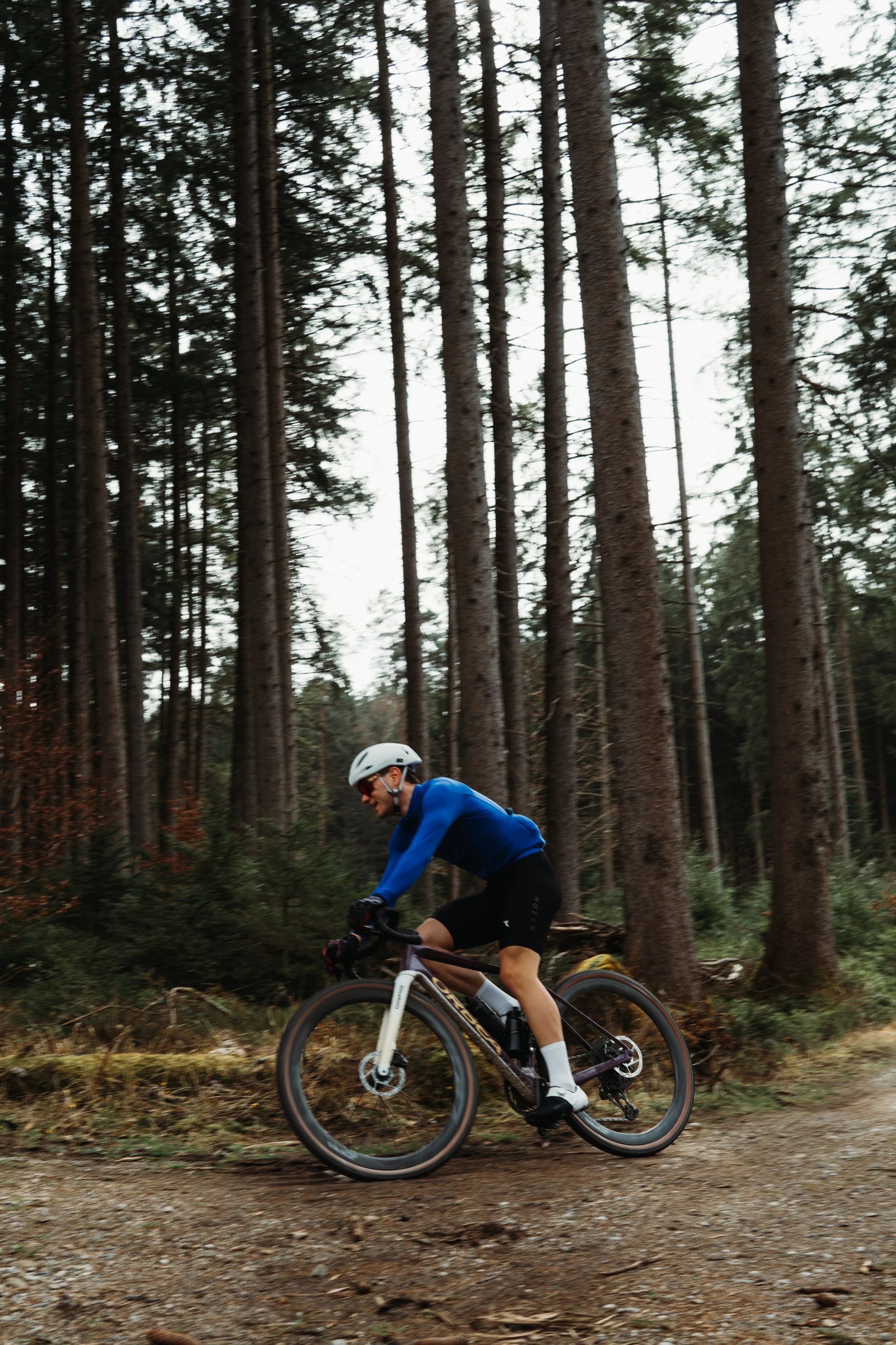 A person is riding a mountain bike through a forest with tall trees. The rider wears a white helmet, a blue jacket, and black shorts. The trail is uneven with dirt and roots visible.