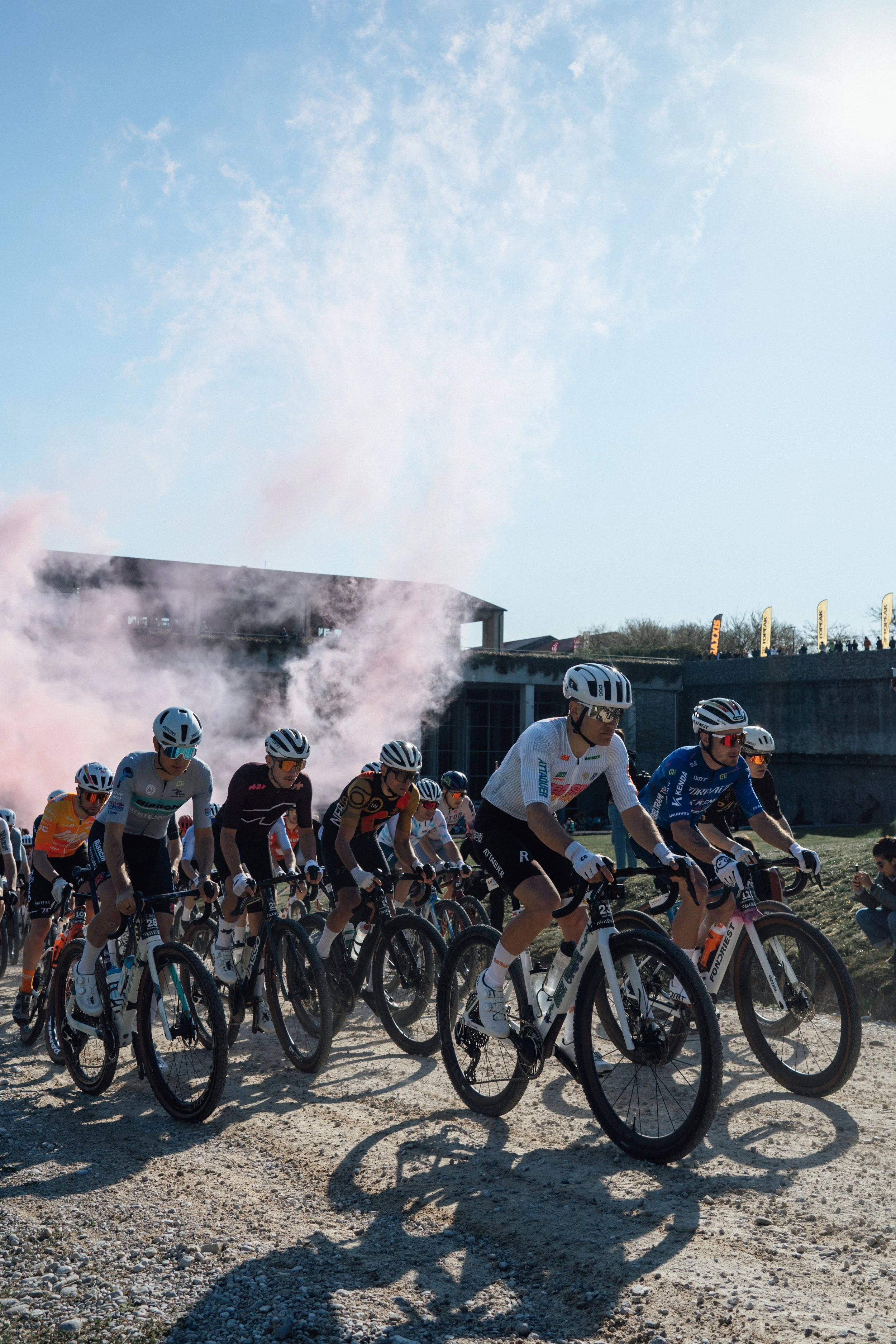 Group of cyclists participating in a race on a dirt trail with buildings and flags in the background under a partly cloudy sky.