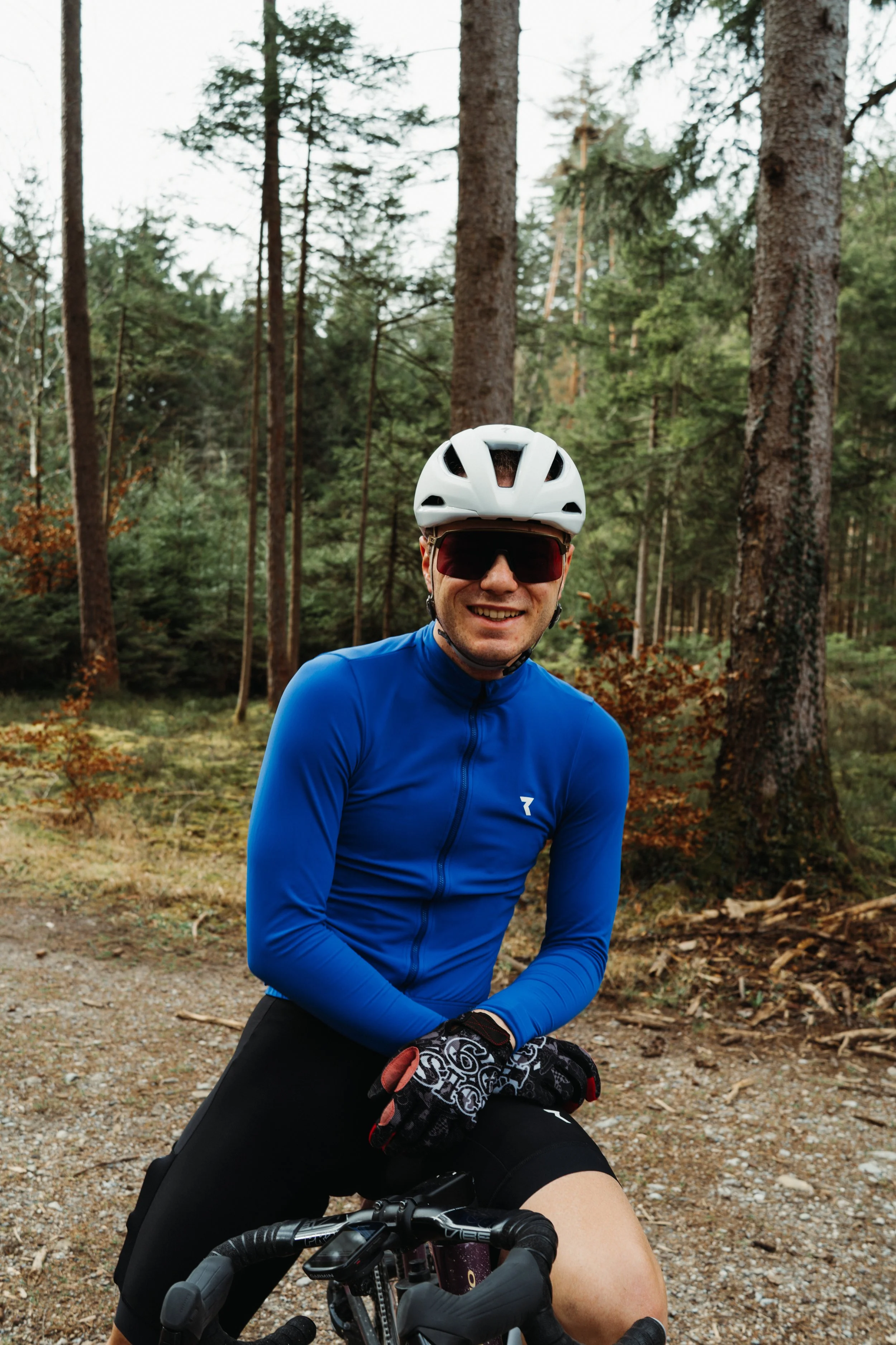 A man wearing a white helmet, sunglasses, and a blue cycling jersey stands outdoors in a forest, smiling at the camera while holding a mountain bike.