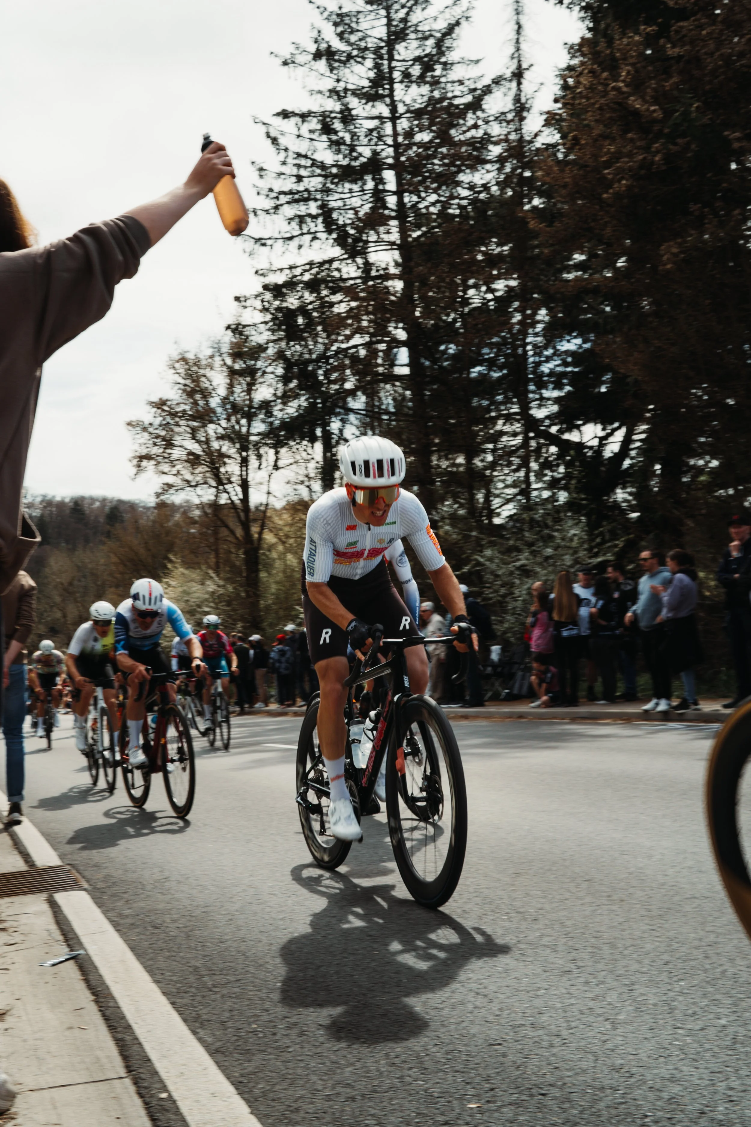 Cyclists compete in a race on a city street with spectators watching from the sidewalk, tall trees line the background on a cloudy day.