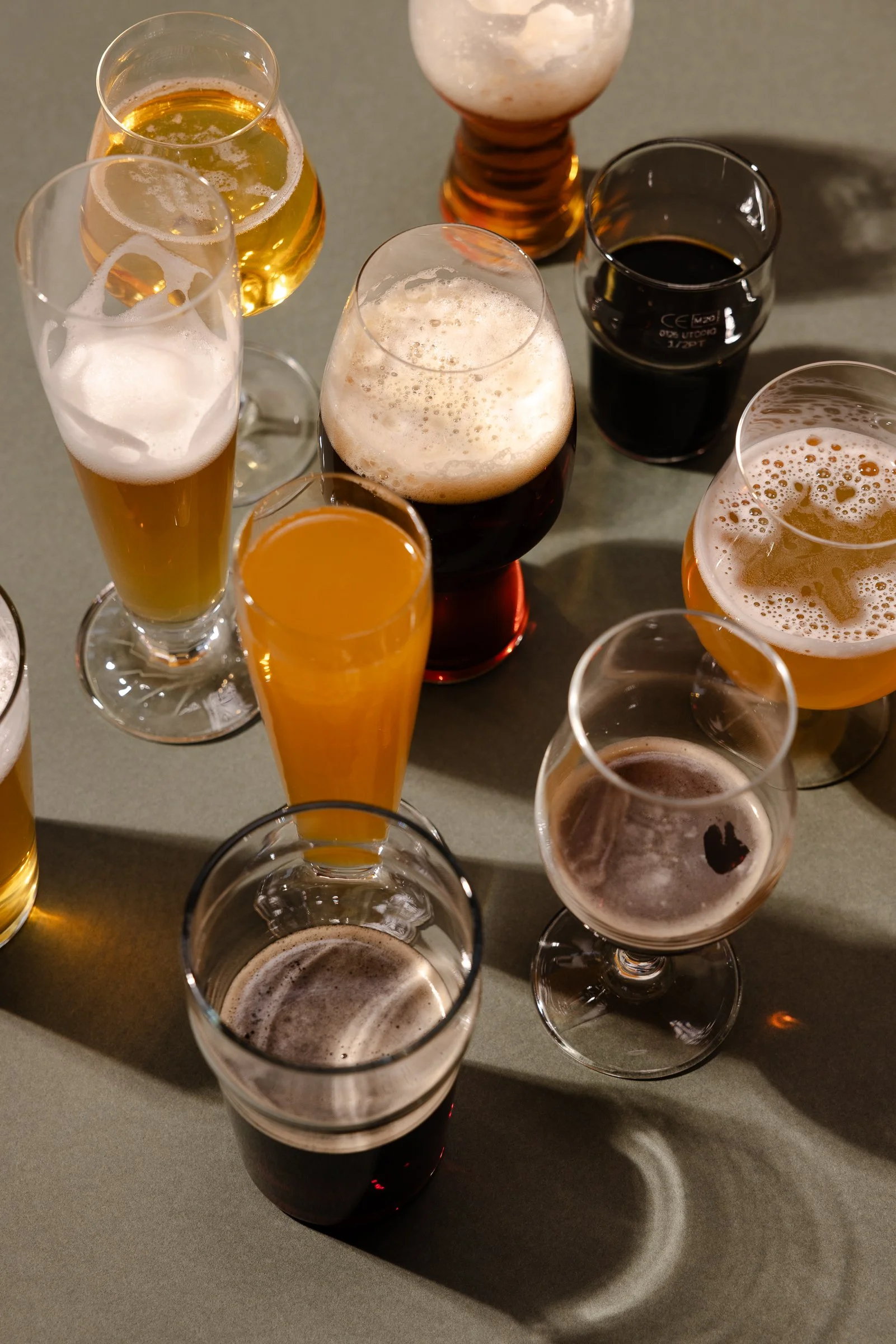 Mehrere Biergläser verschiedener Biersorten, einige mit Schaum, auf einer dunklen Oberfläche, mit Schatten und Lichtreflexen.