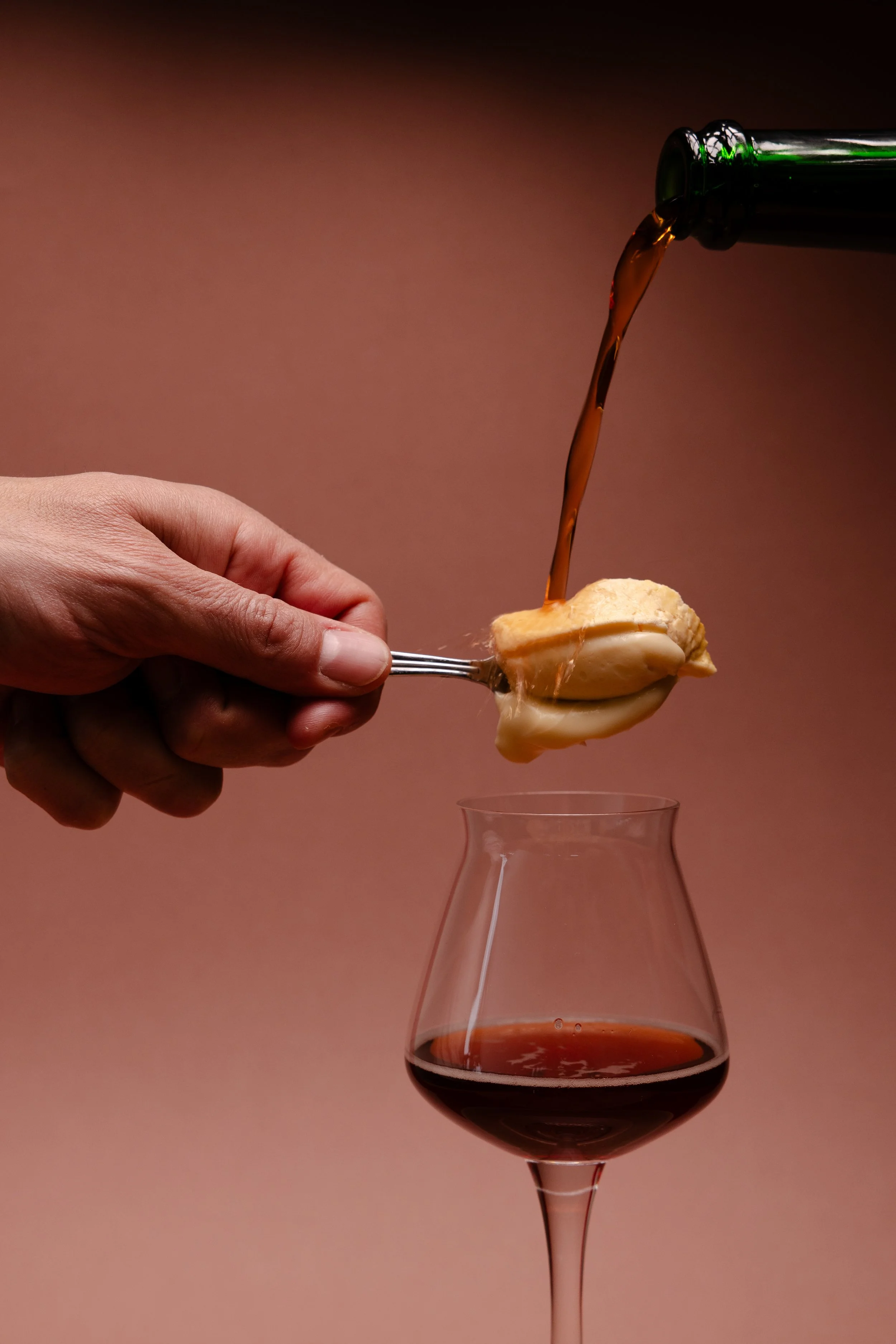 Hand hält einen Löffel mit Weichkäse über einem Glas mit Kriek-Bier welches aus einer Flasche in das Glas gegossen wird.