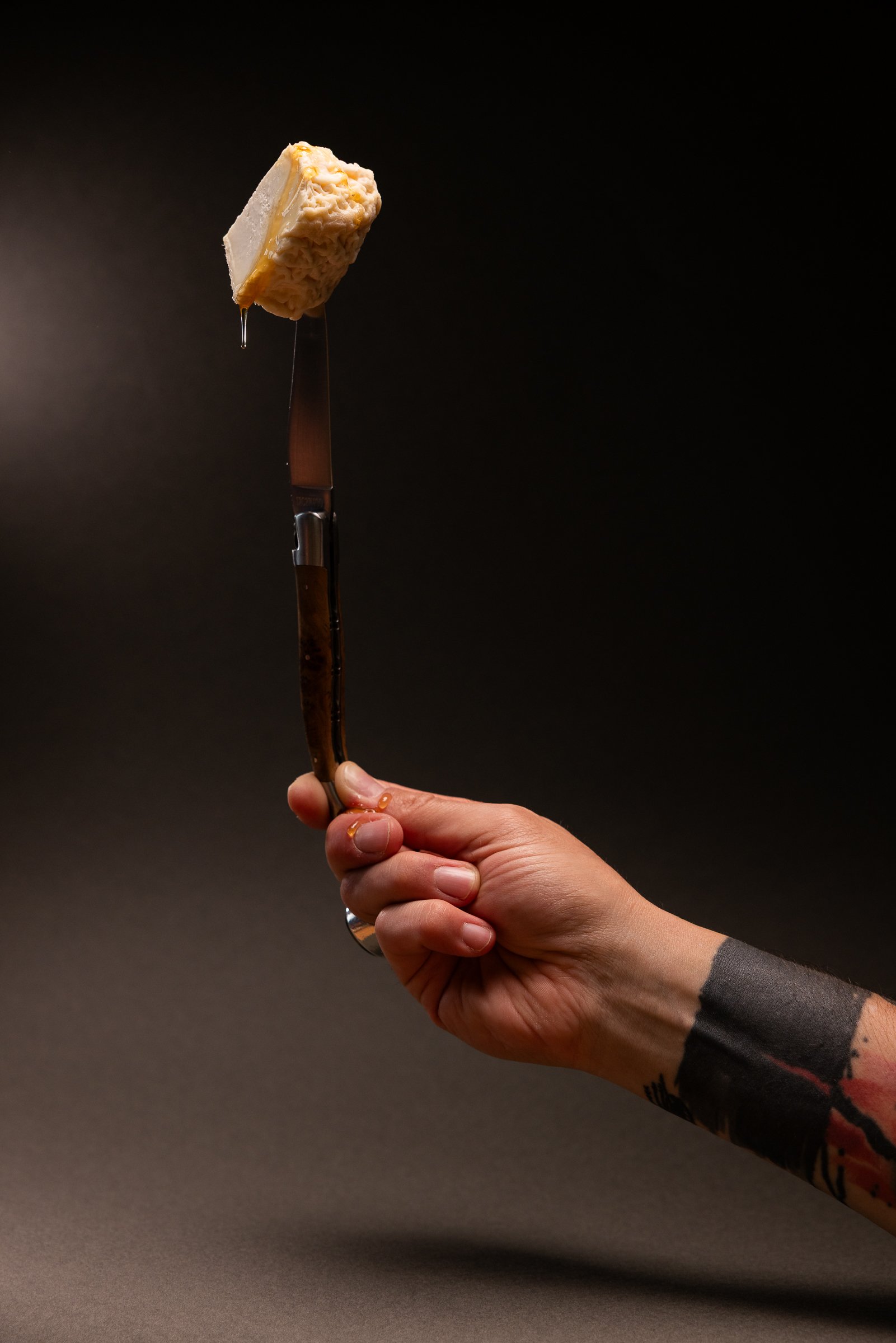 Hand hält ein Messer, unter dem ein Stück Käse hängt, gegen einen dunklen Hintergrund.