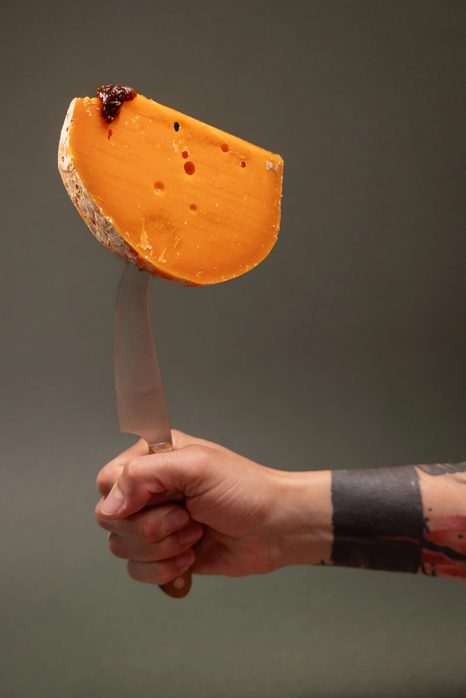 Ein Stück Käse auf einem Messer, gehalten von einer Person mit tätowierten Armen, vor einem dunkelgrünen Hintergrund.