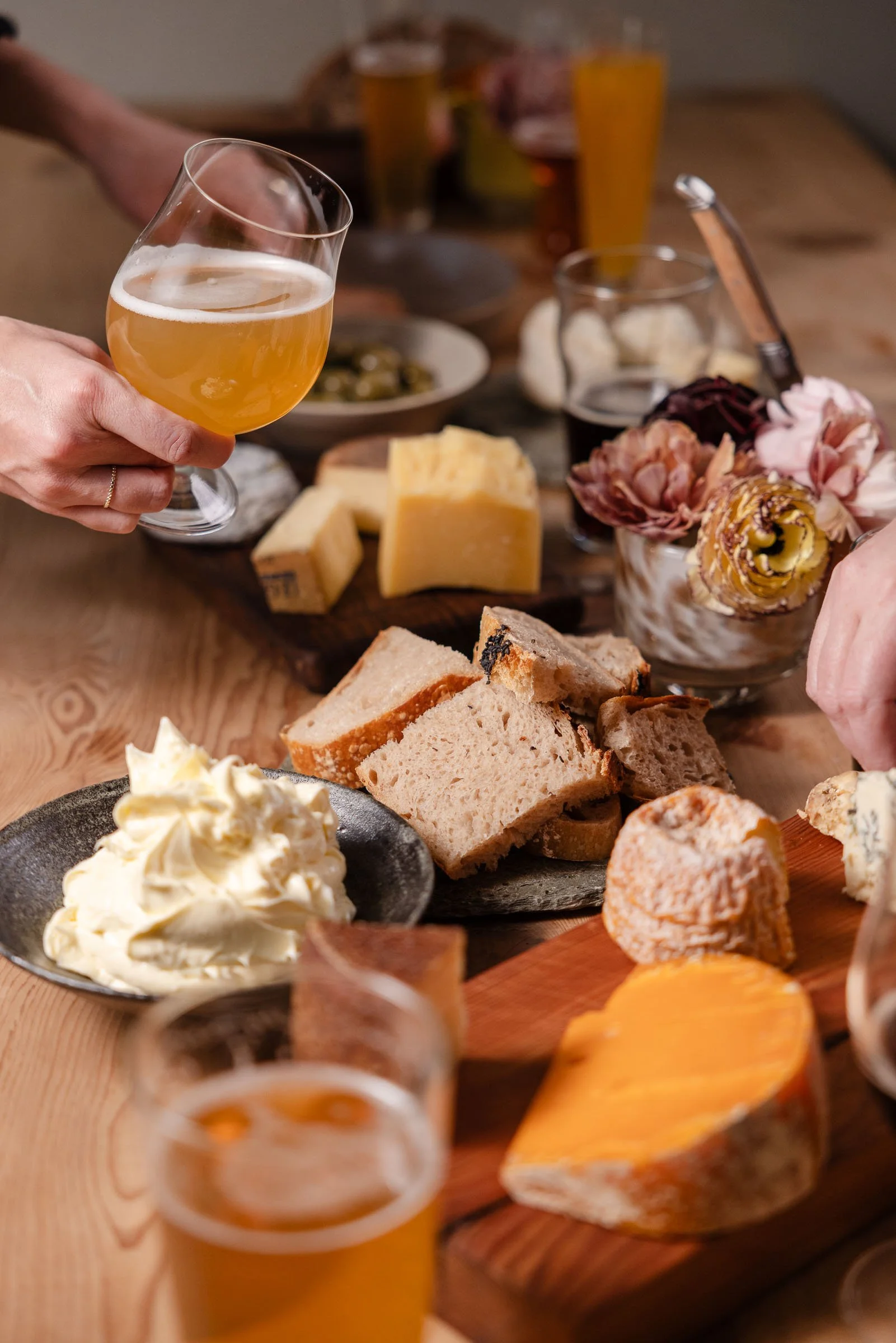 Brett mit Käse, Brot, Wurst und Käseplatte, im Hintergrund Bier, und eine Person hebt ein Glas Bier.