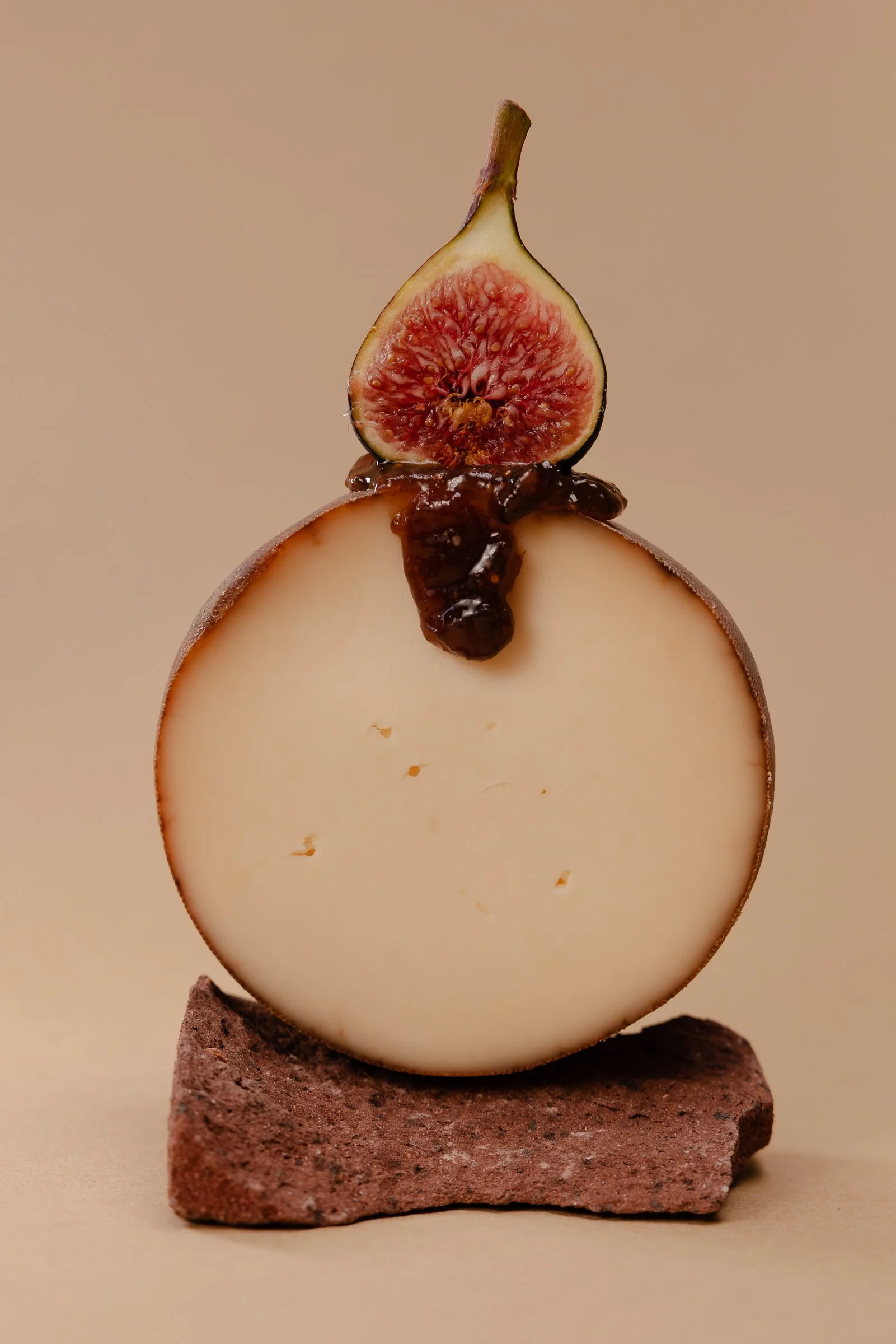 Ein angeschnittener runder Käse, darauf eine halbe Feige mit roten Fruchtfleisch, das ganze liegt auf auf einem braunen Steinblock