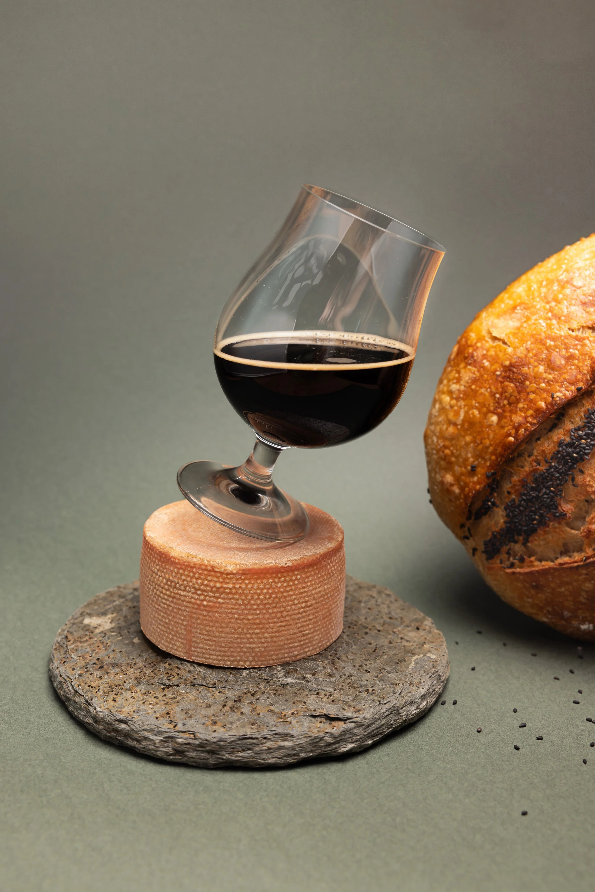 Ein Bierglas mit dunklem Bier steht auf einem Käse, der auf einem runden, steinernen Untergrund liegt. Im Hintergrund befindet sich ein rustikales, rundes Brot.
