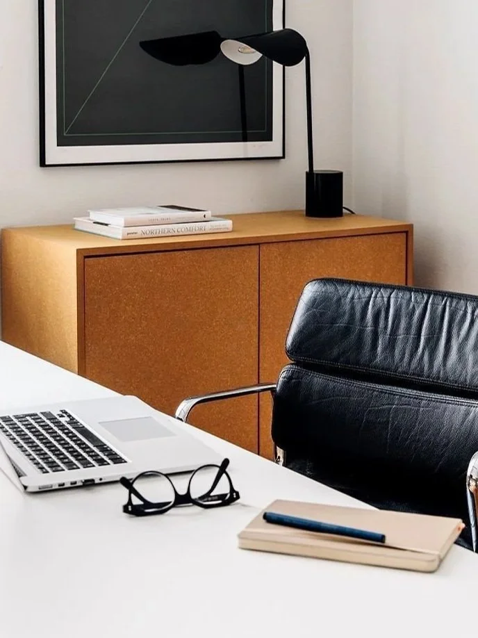 Un bureau avec un ordinateur portable, une paire de lunettes, un cahier avec un stylo, et une chaise en cuir. En arrière-plan, un meuble en bois, une lampe noire, et une œuvre d'art suspendue.