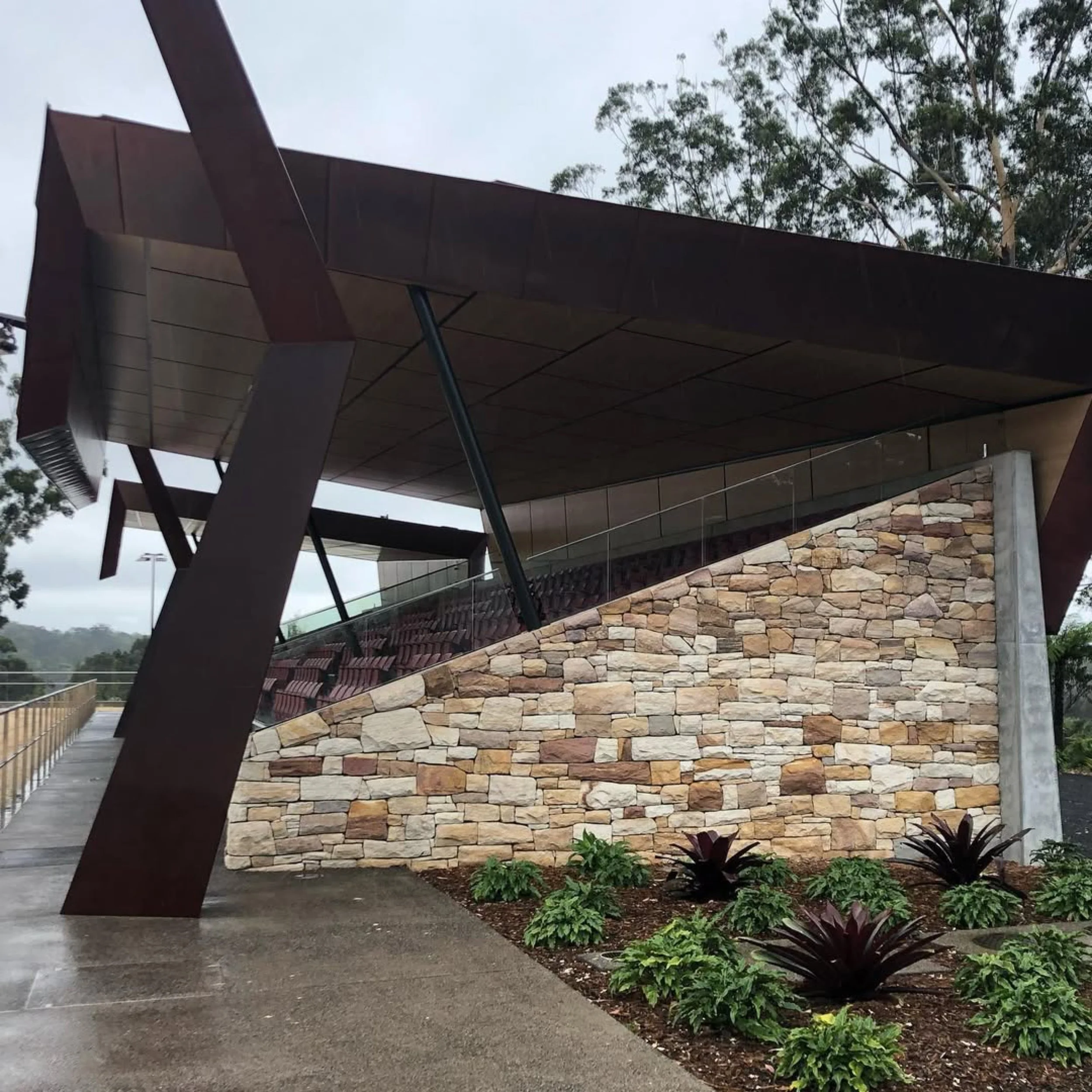 Modern building with a stone facade, dark brown overhanging roof, and landscaped plants at the base