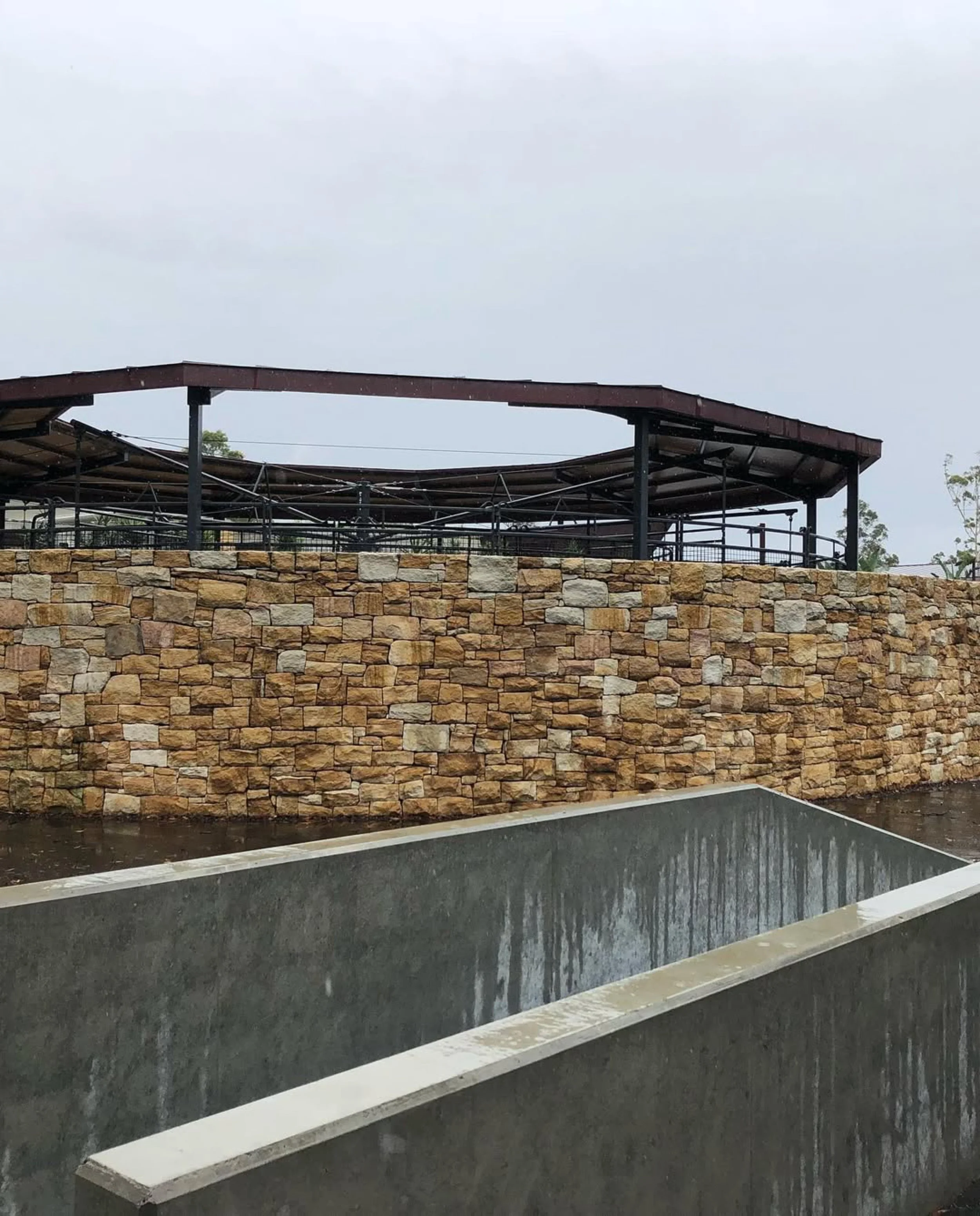 Outdoor scene with a stone wall, a metal railing, and a covered elevated structure, with a concrete water feature in the foreground.